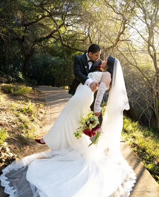 Couple in wedding attire sharing a kiss on a forest path, woman holding a bouquet of flowers, surrounded by trees and sunlight.
