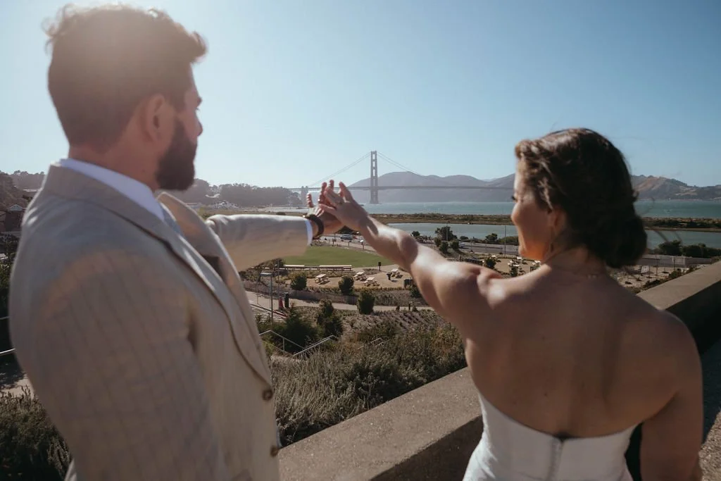 A couple stands on a hilltop during sunset, reaching out to hold hands with the Golden Gate Bridge and San Francisco skyline visible in the background.