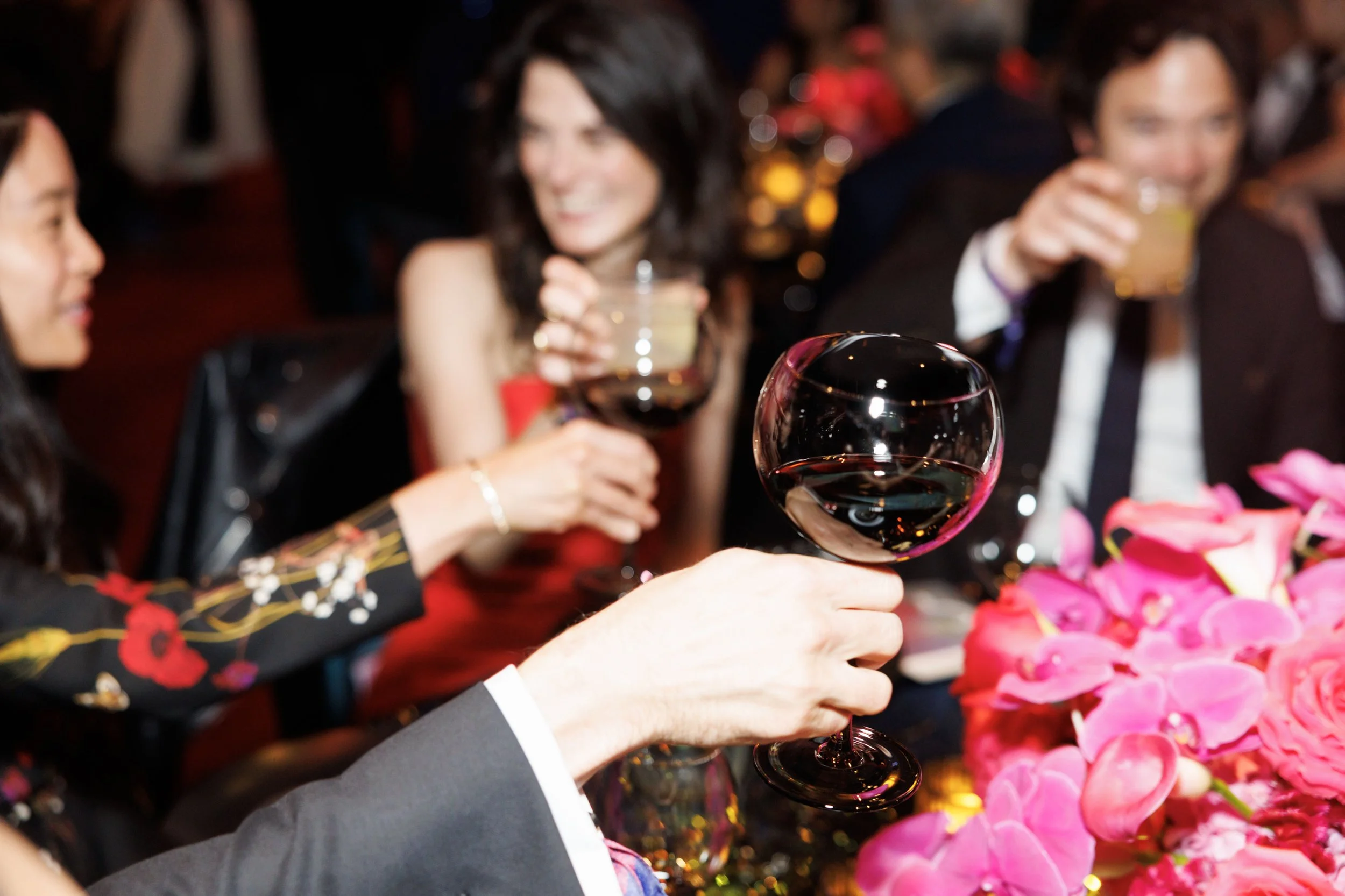 People toasting with glasses of red wine at a celebration table decorated with pink flowers.