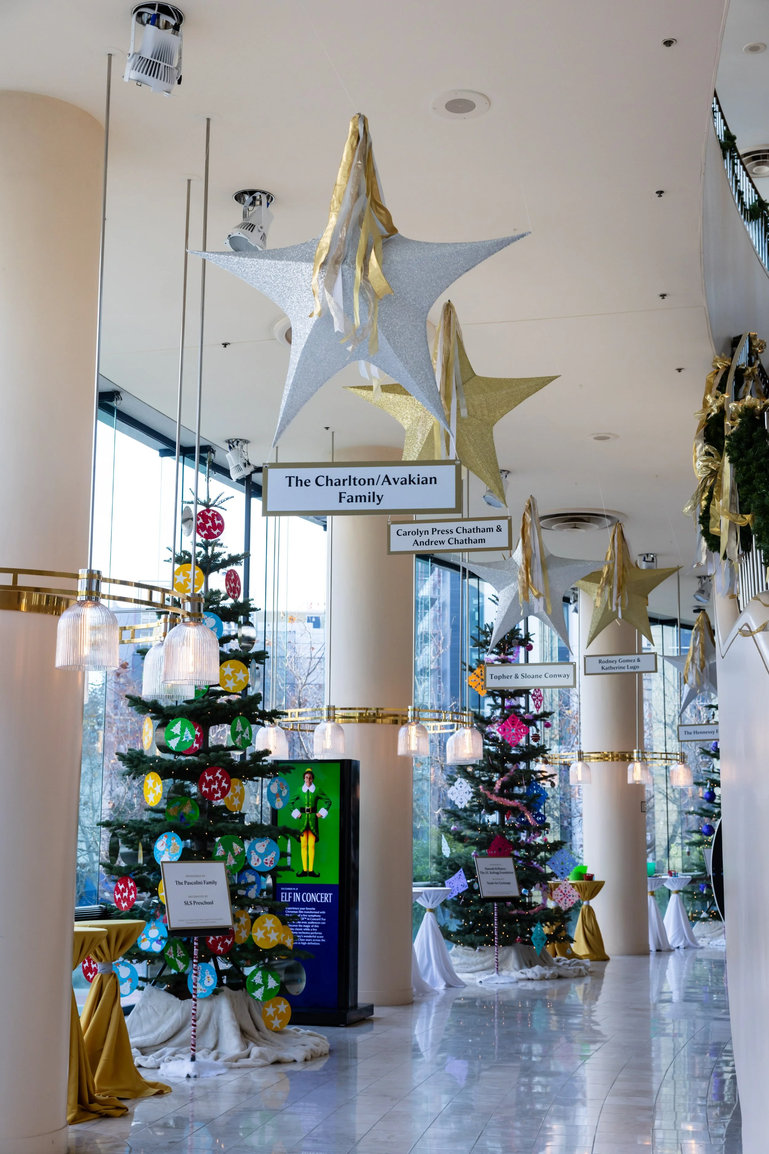 Decorated Christmas trees with ornaments and signs hanging from the ceiling, inside a well-lit building with large windows.