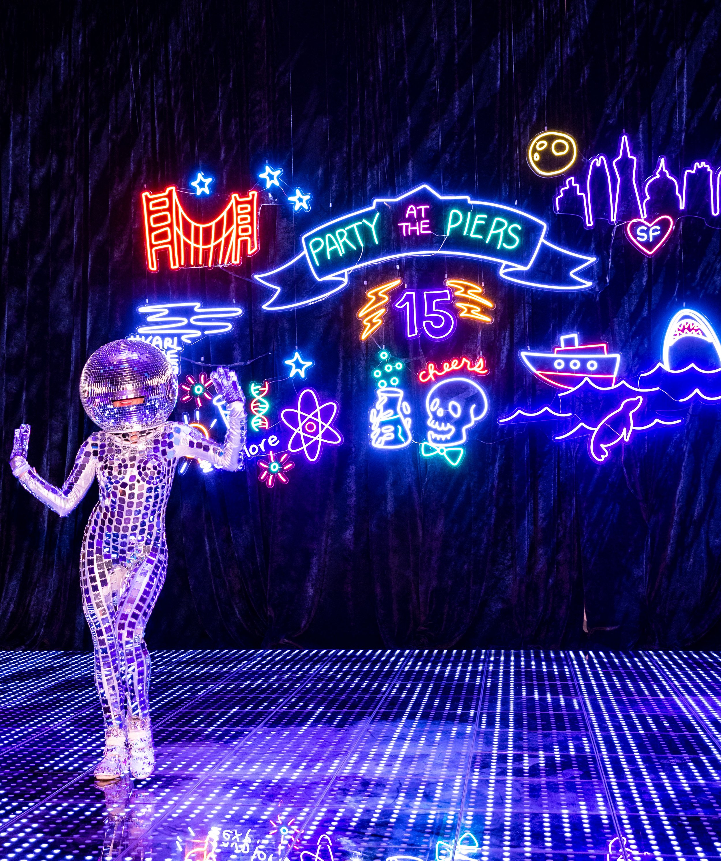 A person in a disco mirror suit with a disco ball for a head standing in front of a neon sign at a party. The neon sign includes various colorful designs and text such as "Party at the Piers 15," a city skyline, a boat, a whale, a skull with a beaker, and additional decorative elements.