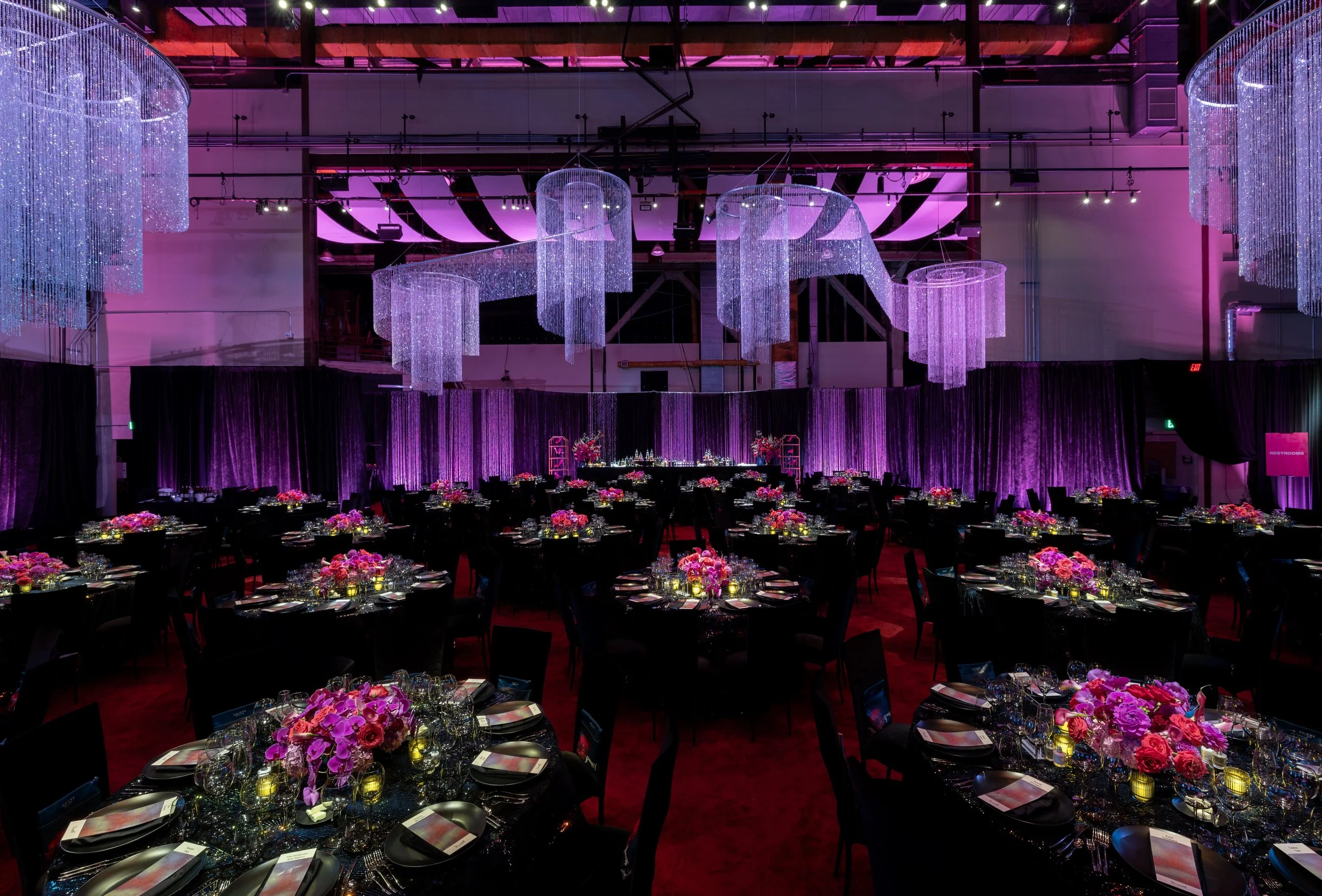 Elegant banquet hall decorated with pink and purple lighting, featuring round tables with black tablecloths, floral centerpieces, and place settings, with large hanging crystal chandeliers.