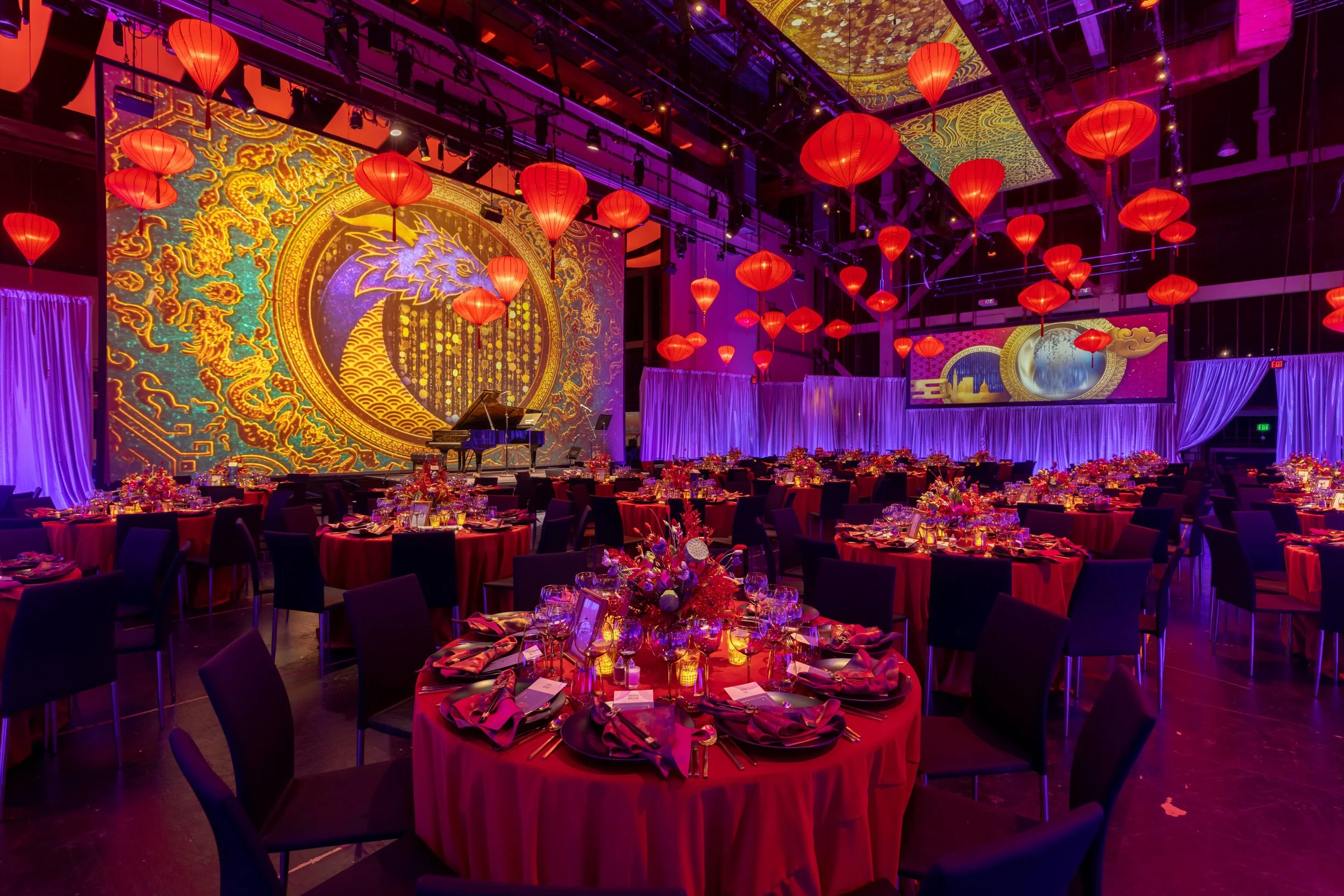 A decorated event hall with red lanterns hanging from the ceiling, round tables with red tablecloths, and centerpieces. The background features a large projection of a dragon and moon in gold and purple hues, with purple curtains and additional screens displaying similar patterns.