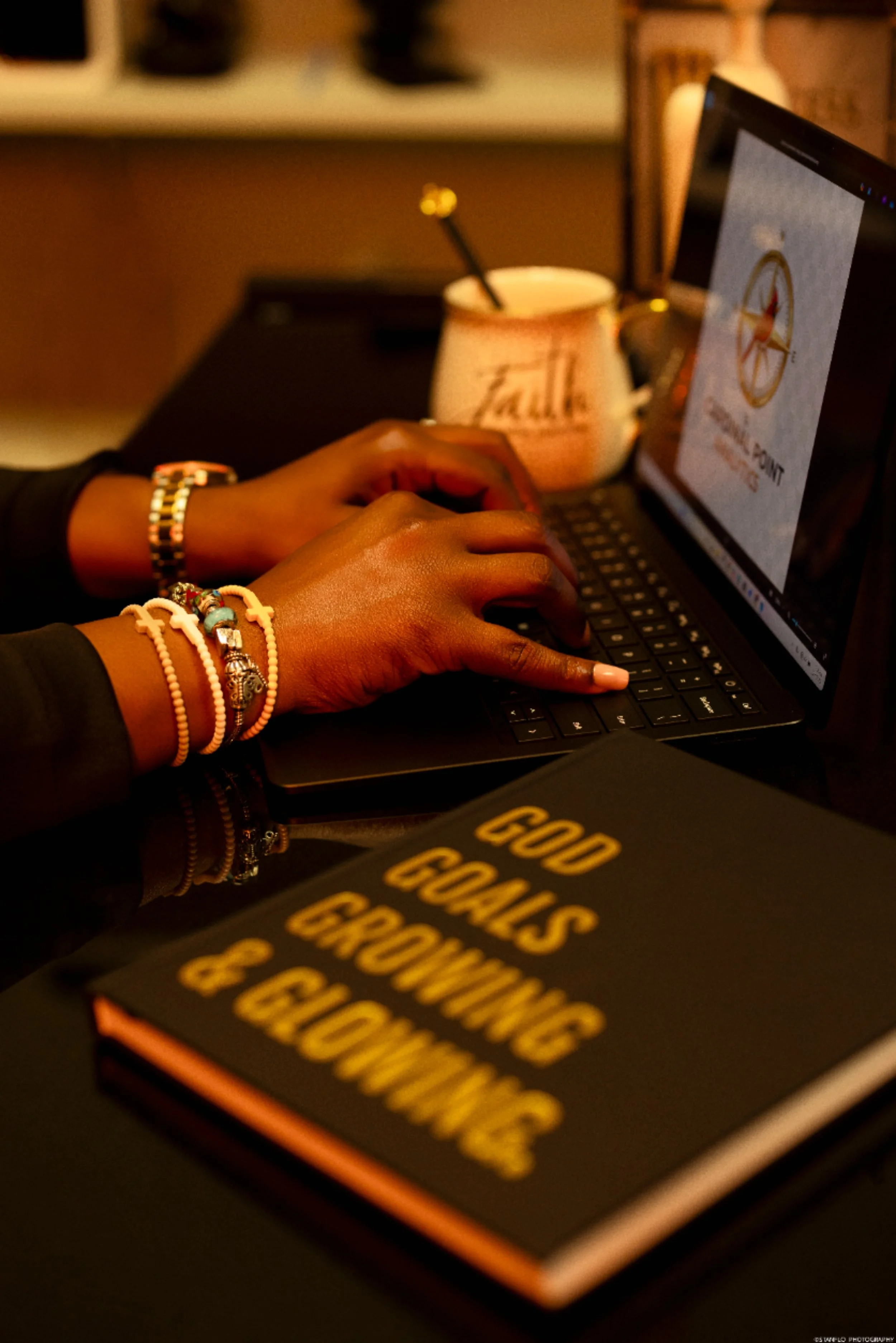 Person typing on a laptop with a notebook titled "GOD GOALS GROWTH & GLOWING" on a desk, a mug and a computer screen in the background.