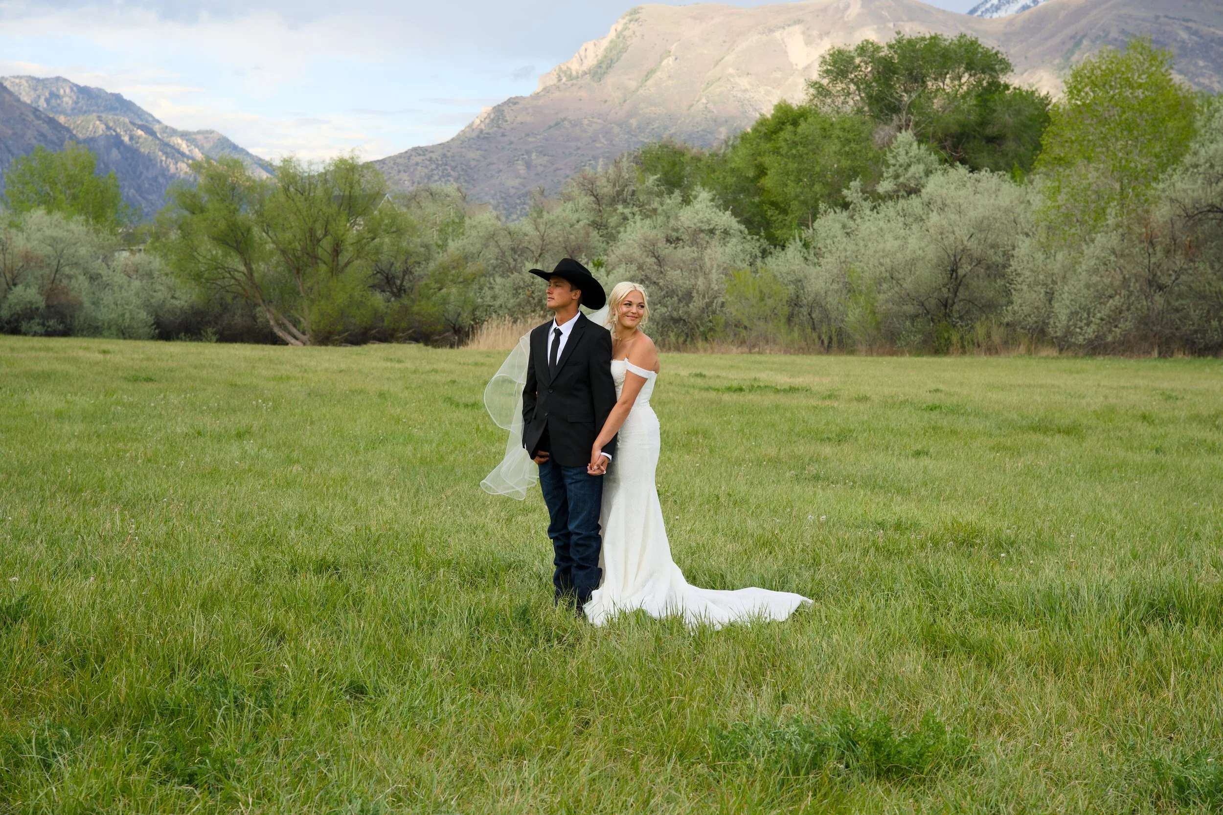 A bride and groom standing in a grassy field with mountains and trees in the background. The groom is wearing a black suit, white shirt, tie, and a black hat, while the bride is in a white off-shoulder wedding dress with a veil. They are holding hands and facing different directions, with the bride smiling.