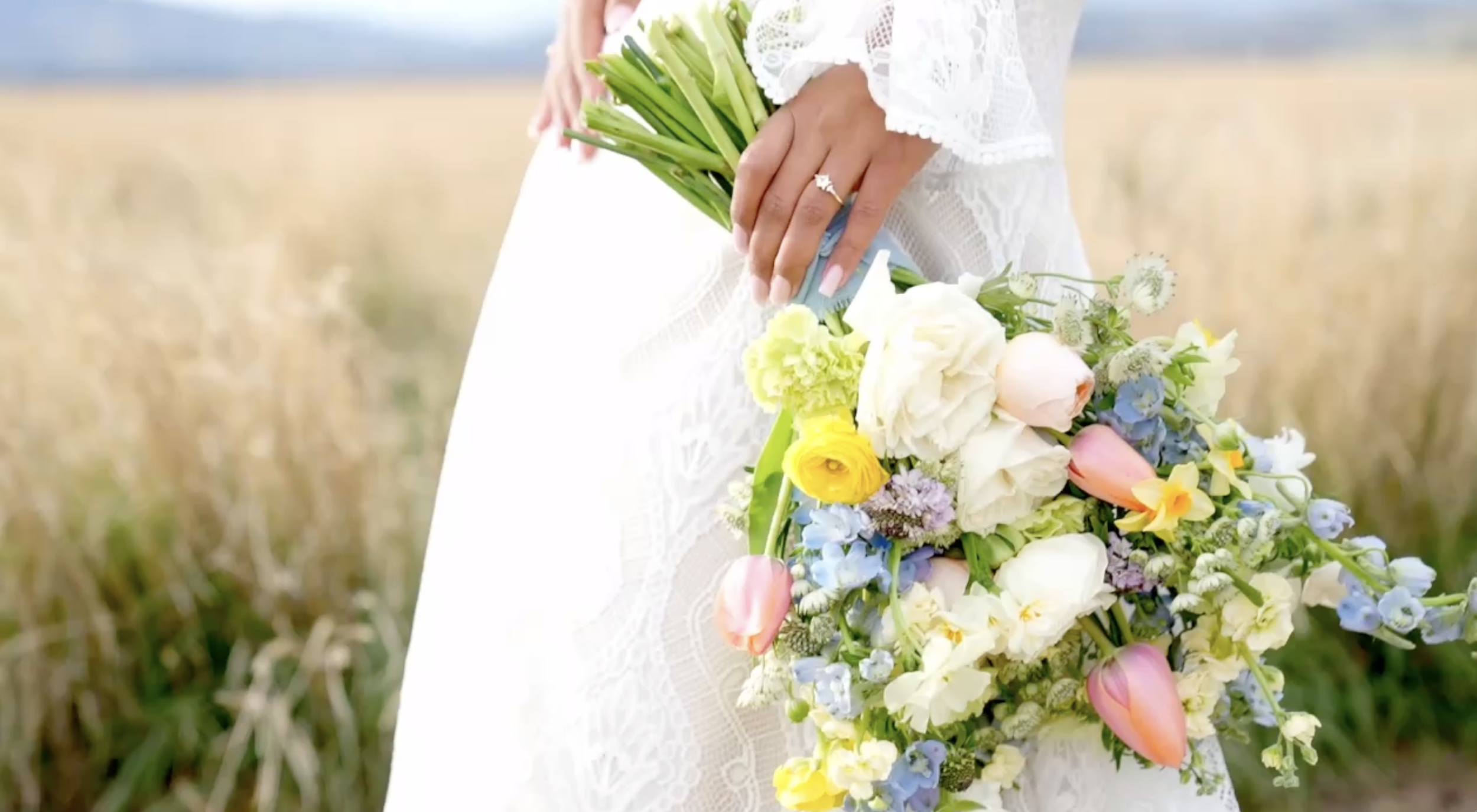 A woman wearing a white lace dress holding a large bouquet of colorful flowers outdoors, with a blurred field in the background.