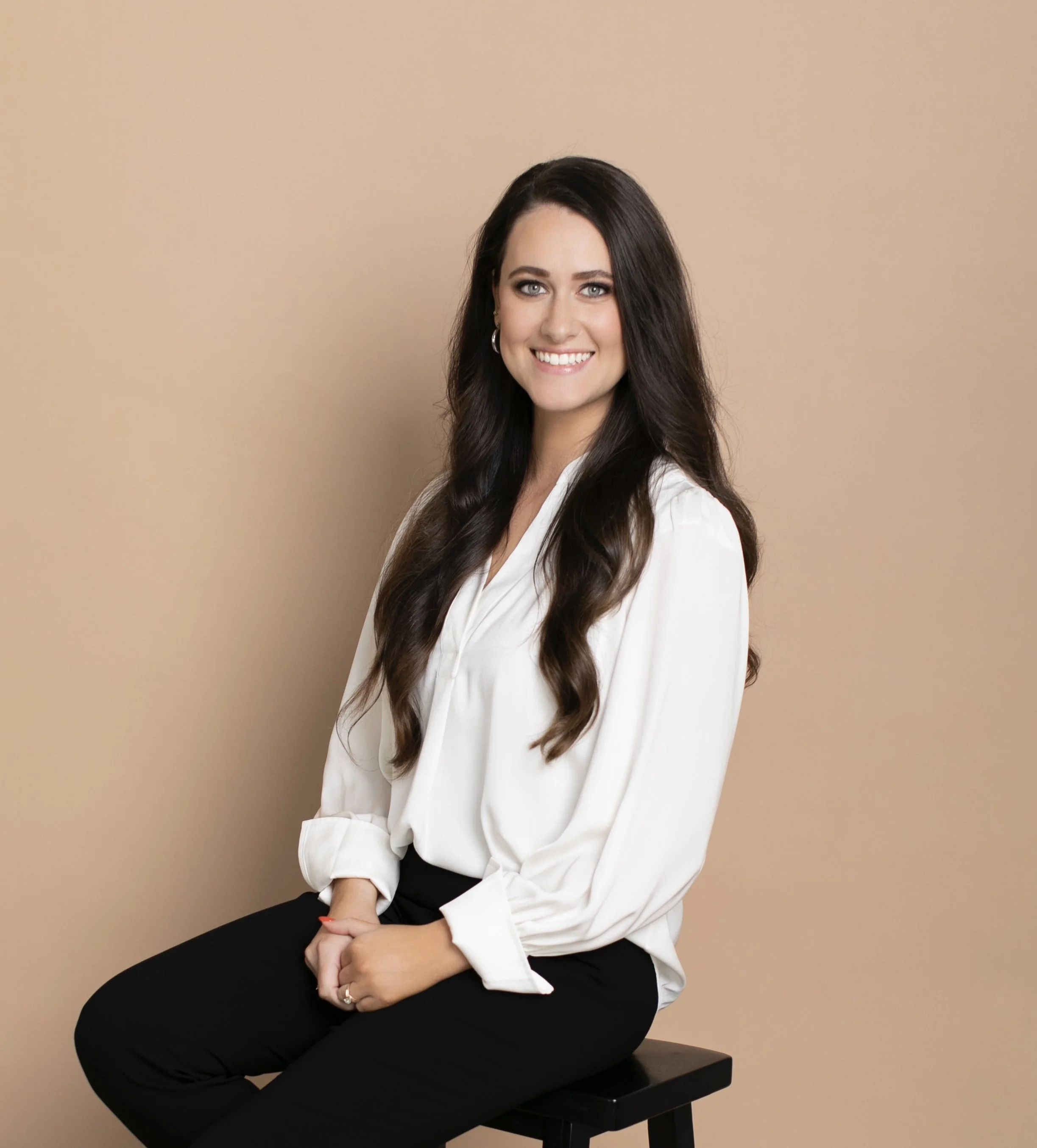 A woman with long dark hair and blue eyes, sitting on a black stool against a beige background, smiling at the camera. She is wearing a white blouse and black pants, with her hands resting on her lap.