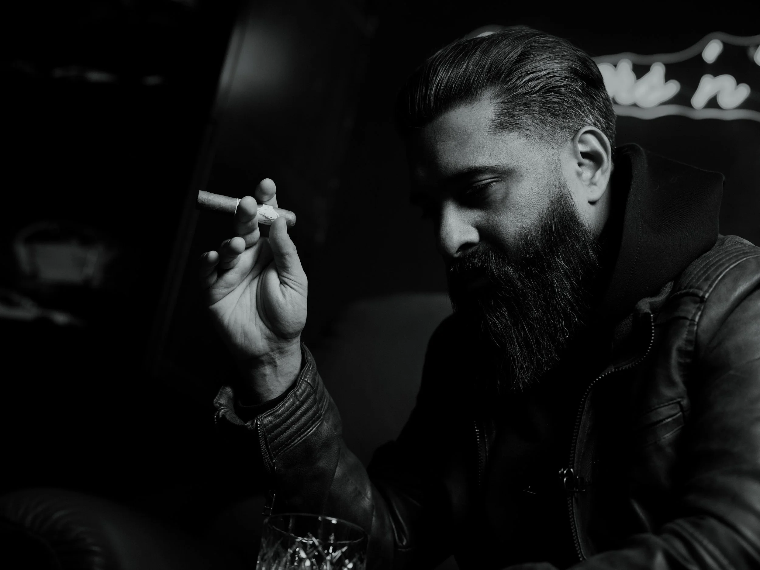 A man with a beard, slicked-back hair, and wearing a leather jacket, holding a cigarette near his face in a dimly lit setting.
