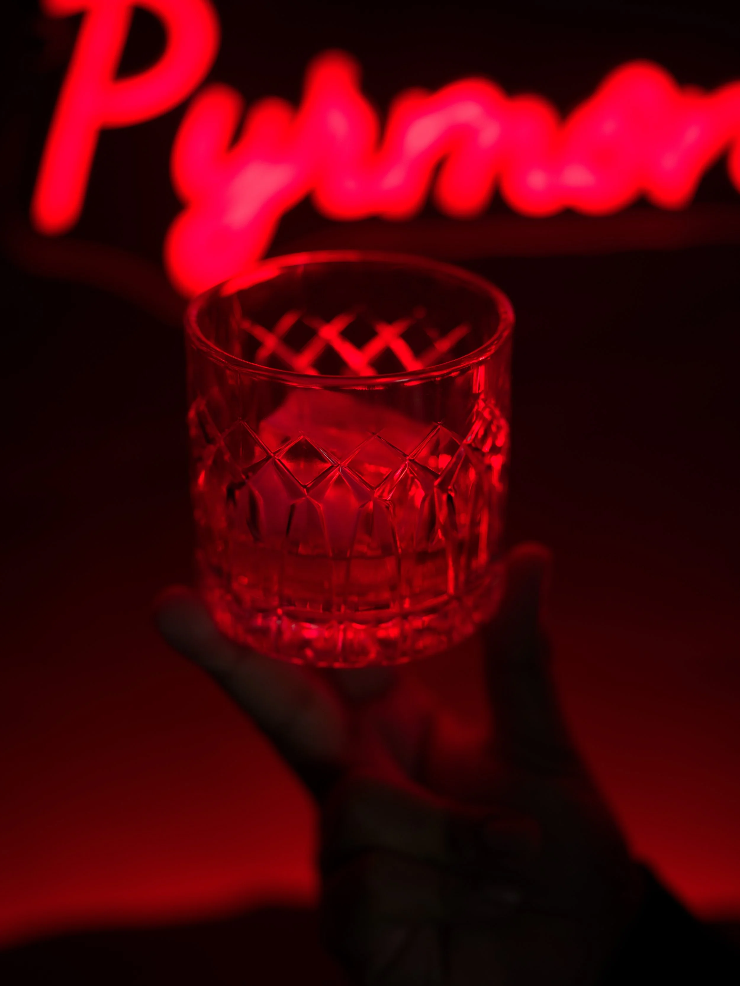 A person holding a glass with a diamond pattern, illuminated by red light, with a red neon sign in the background that says 'Pyramid'.
