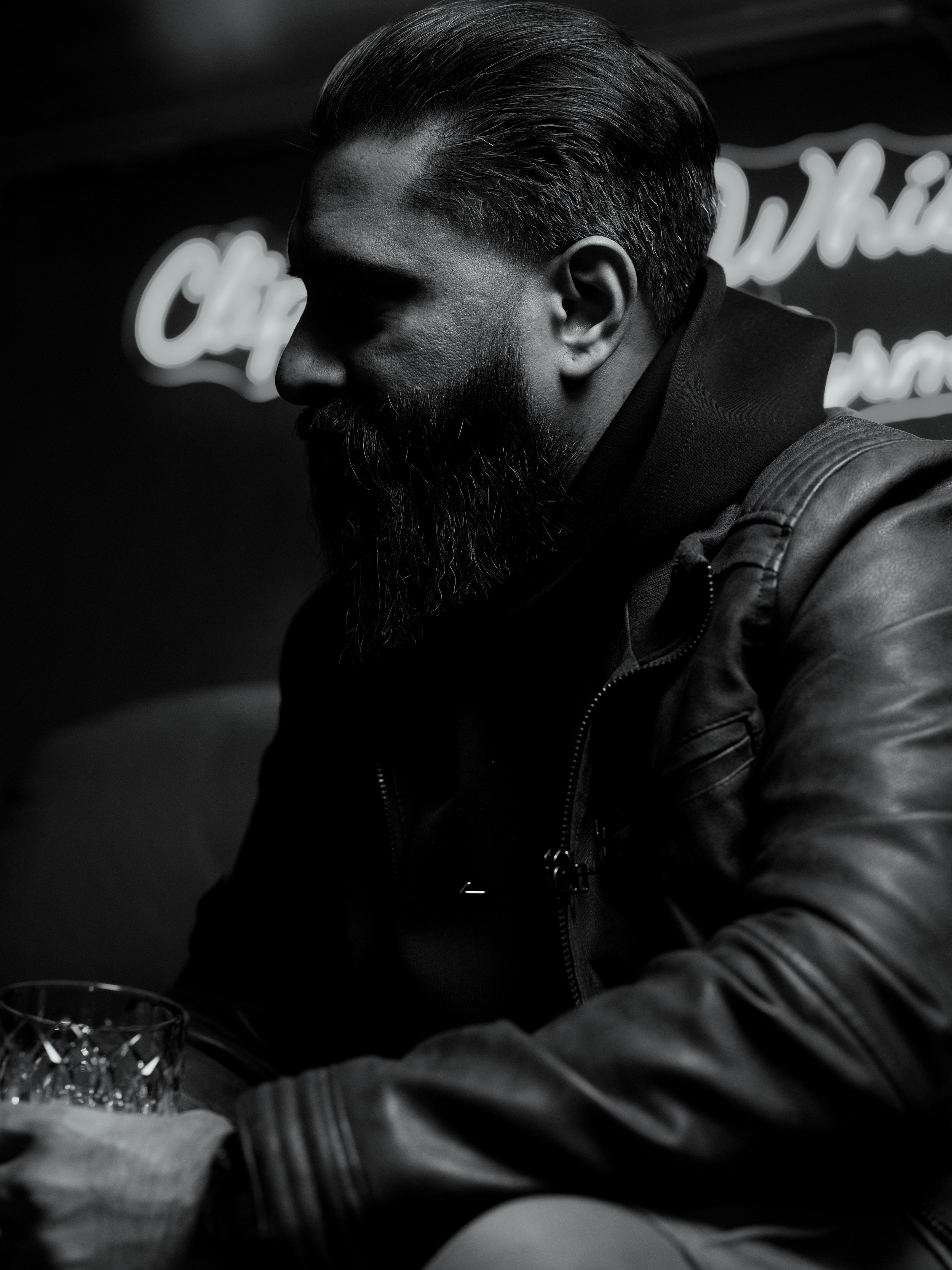 Black and white photo of a bearded man with slicked back hair wearing a leather jacket, seated with his head tilted downward, with a glass in front of him.