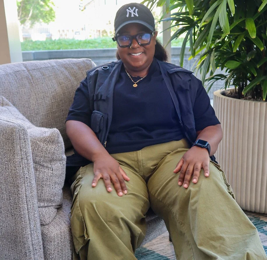 Woman sitting on a beige sofa, wearing a black NY cap, sunglasses, a black shirt, khaki cargo pants, and a black vest, smiling with greenery and large potted plant in the background.
