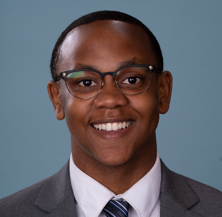 A young man in a suit and tie, wearing glasses, smiling against a plain blue background.