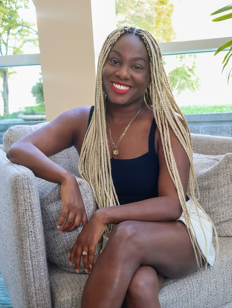 A smiling woman with long blonde braids, wearing a black sleeveless top and white shorts, sitting on a beige sofa near large windows letting in natural light.