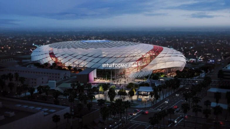 Aerial view of the new Intuit Dome basketball arena at dusk, surrounded by palm trees and city streets.