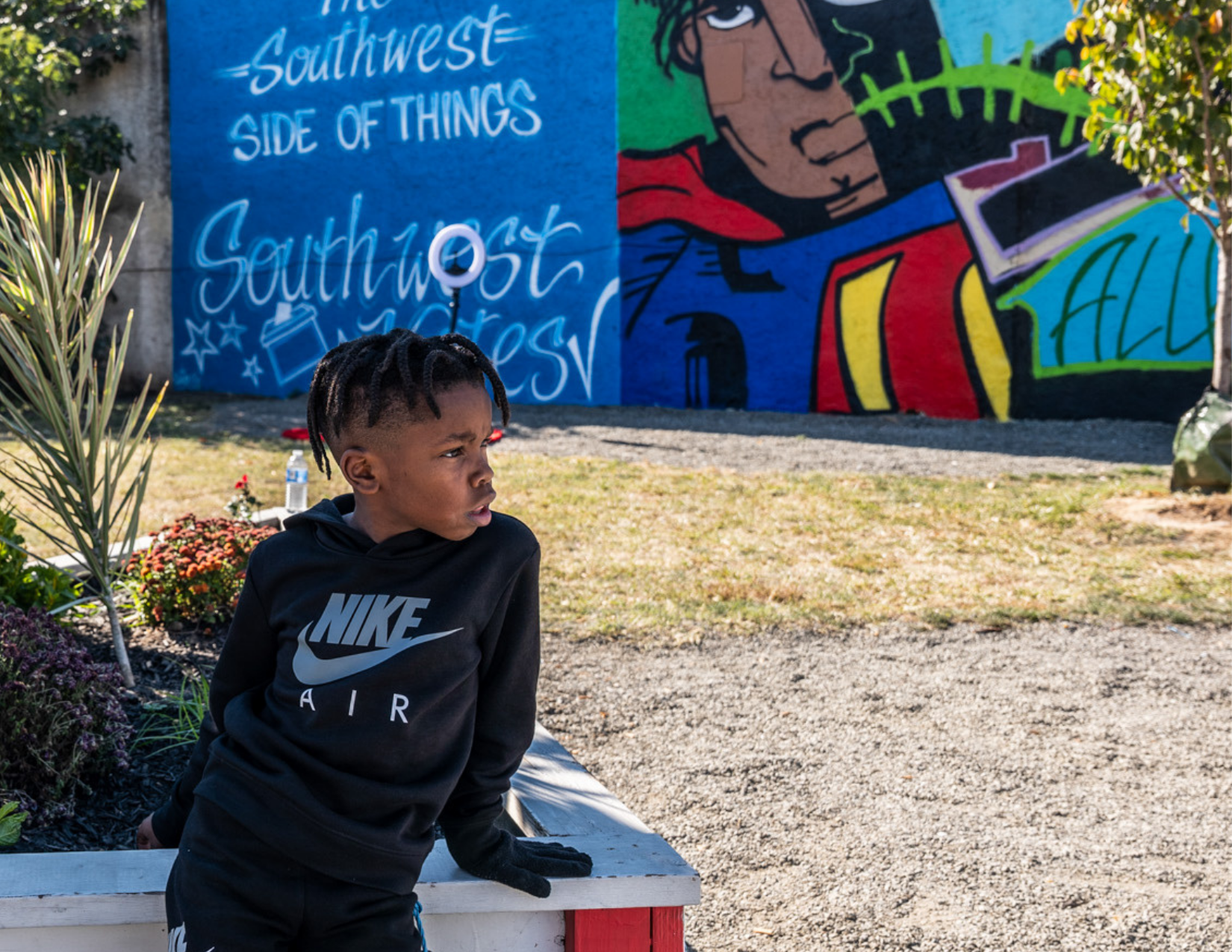 A young boy with dreadlocks, wearing a black Nike Air hoodie, leans on a flower bed in front of a colorful mural on a wall. The mural features abstract figures and text including "Southwest SIDE OF THINGS" and a stylized face.