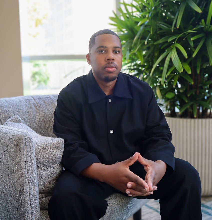 A young man with short hair and a beard, wearing a black shirt, sitting on a gray sofa in a room with a large green plant behind him.