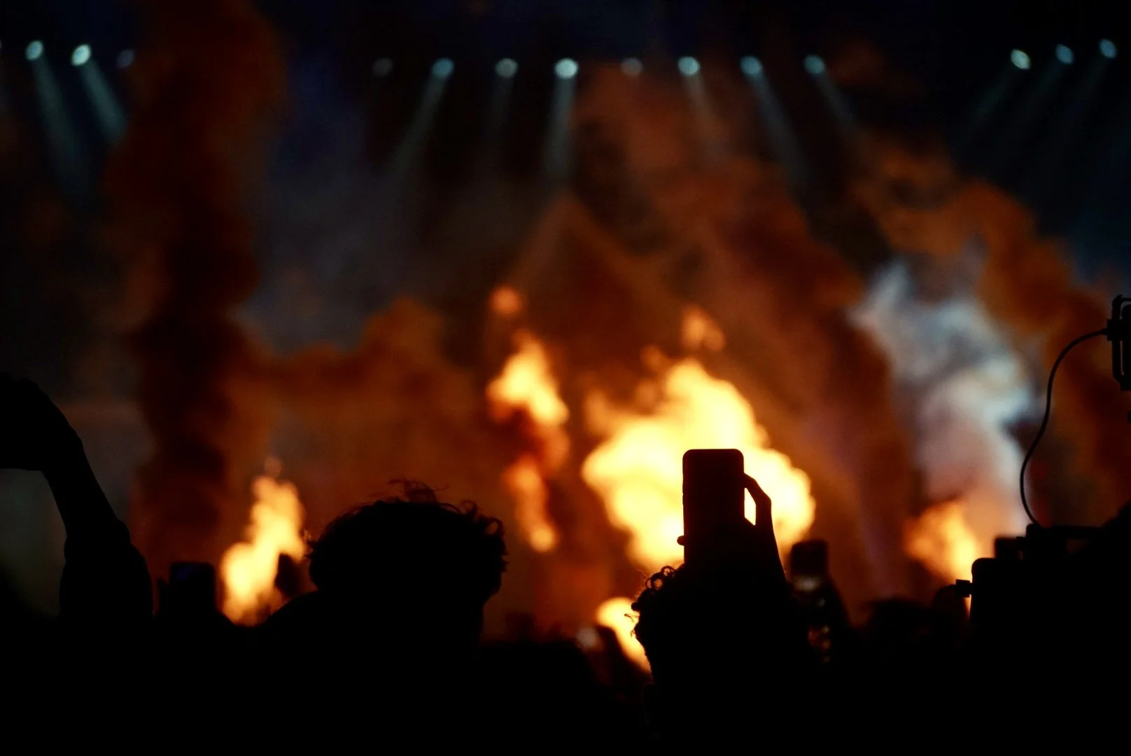 A concert scene with fire and smoke on stage, silhouetted audience members are taking photos.