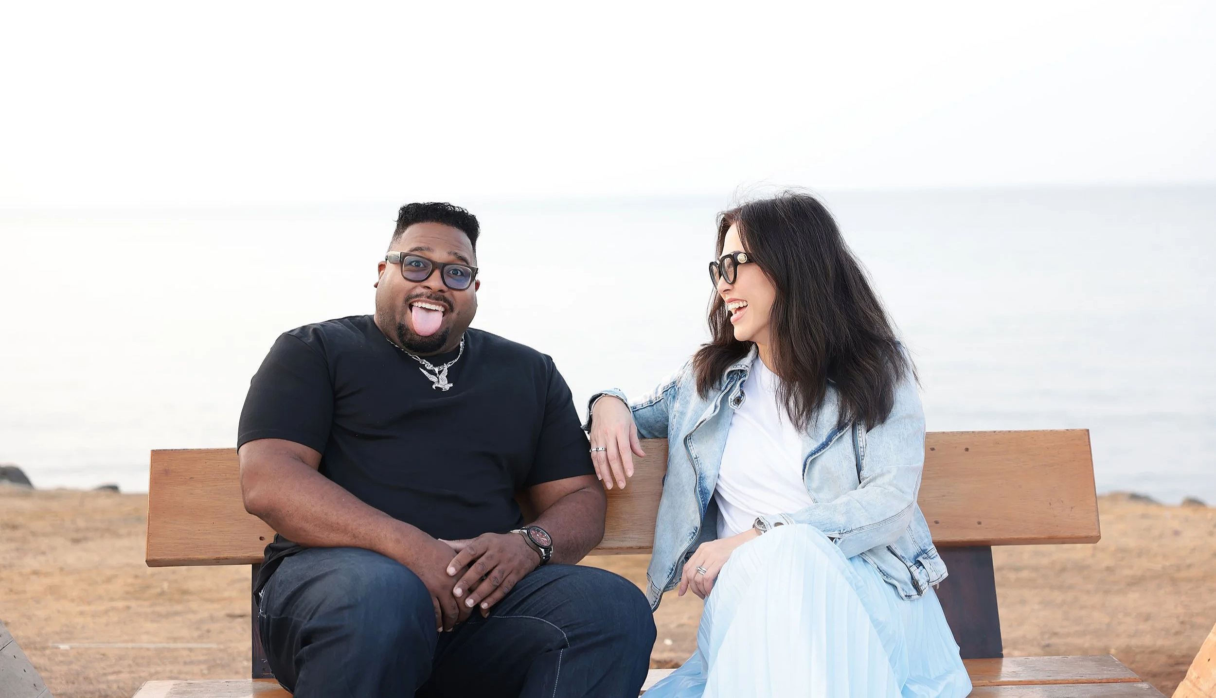 A man and woman sitting on a wooden bench near the beach, smiling and laughing together. The man is wearing a black T-shirt, glasses, and jewelry, and sticking out his tongue. The woman is wearing a light denim jacket, glasses, and a white dress, with her arm resting on the man's shoulder.
