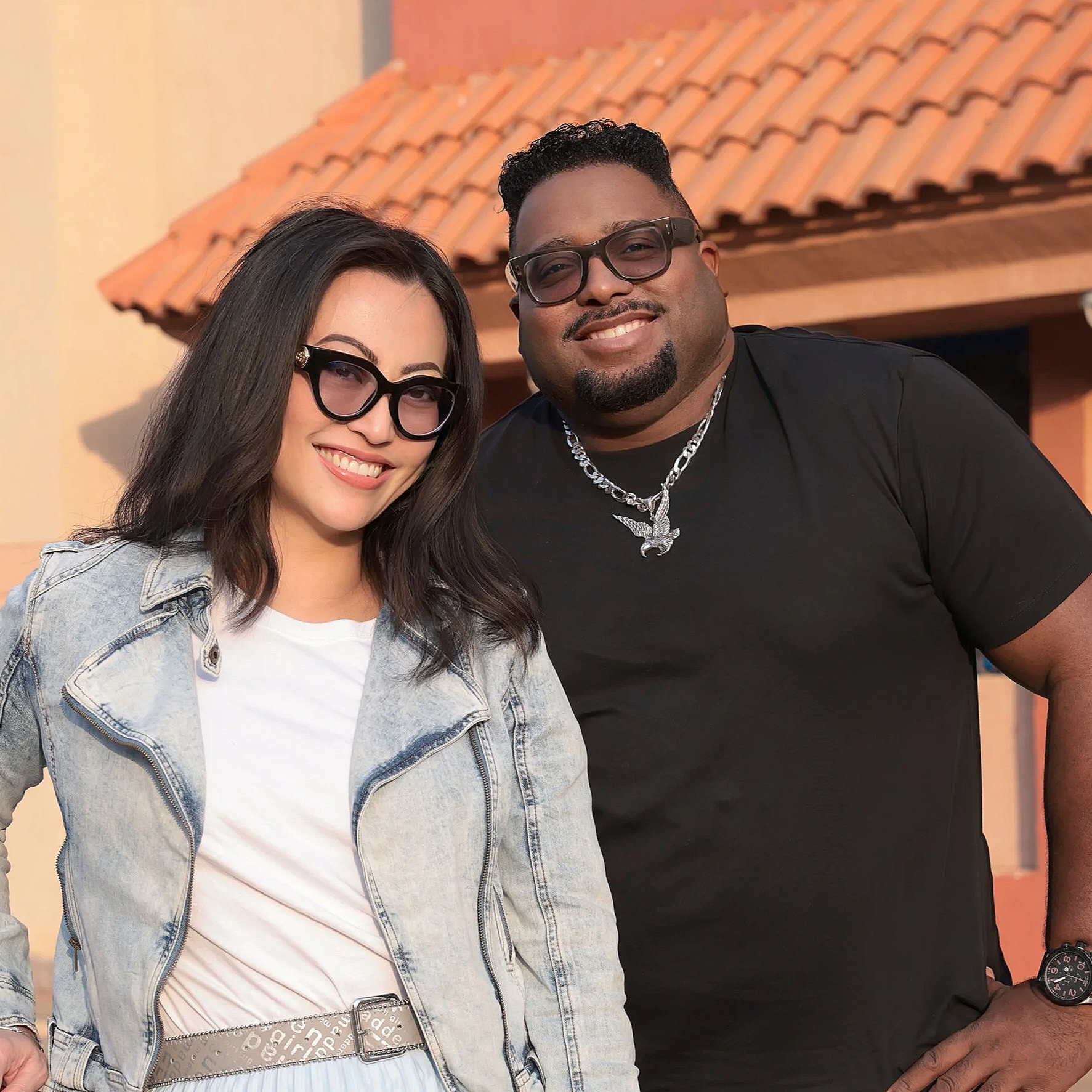 A smiling woman and man stand together outdoors in front of a building with a tiled roof. The woman has long dark hair, glasses, a white t-shirt, and a light denim jacket. The man has short dark hair, glasses, a black t-shirt, a silver chain necklace, and a watch.