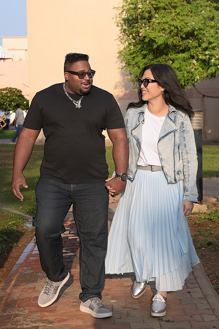 A couple walking hand-in-hand outdoors, middle-aged man and woman, smiling, dressed casually, with trees and a building in the background.