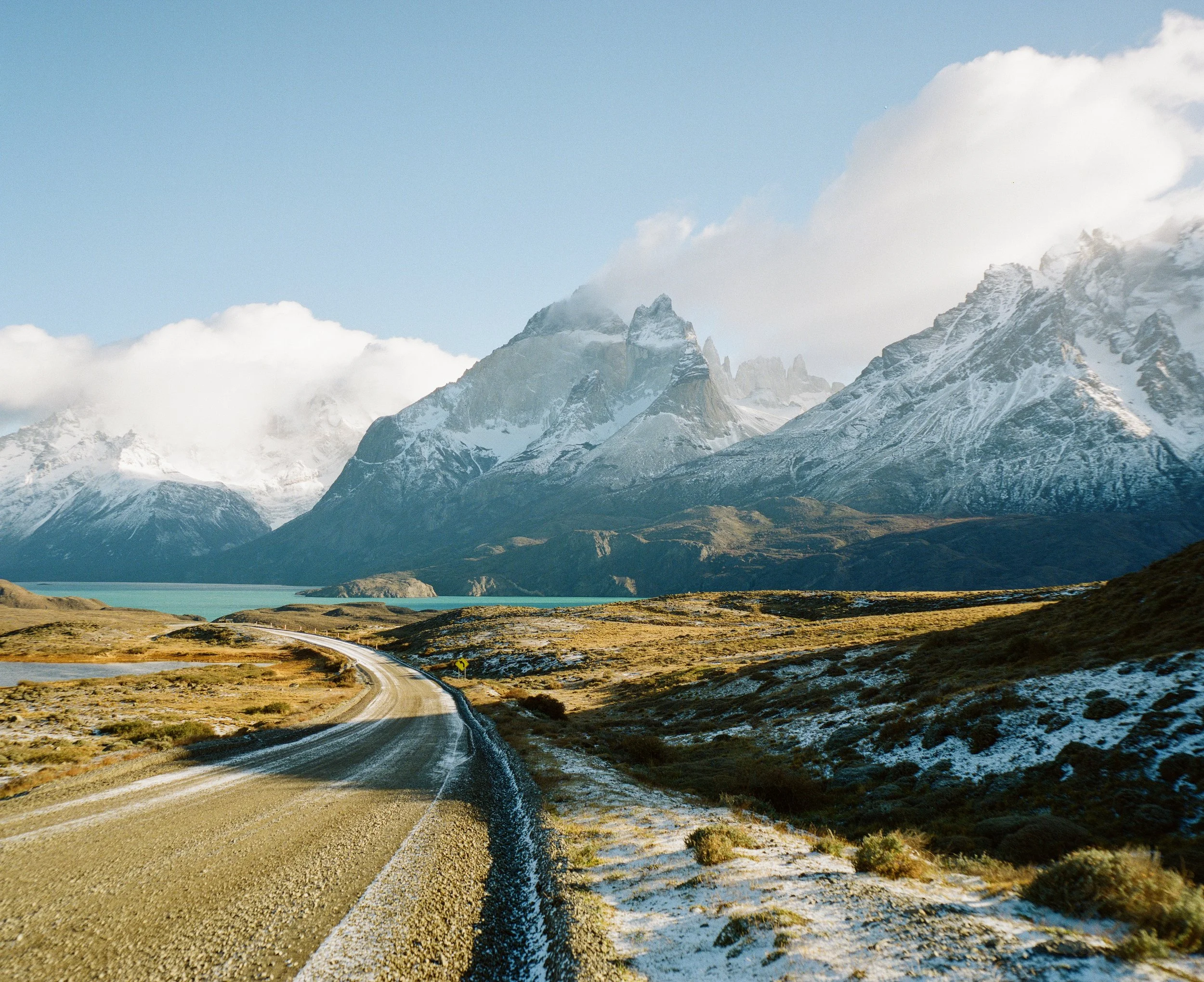 A winding gravel road in a mountainous landscape with snow-capped peaks and a partly cloudy sky.