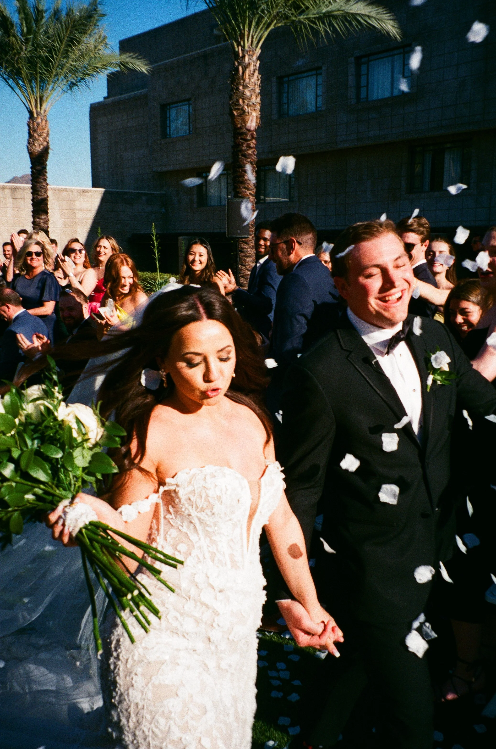 A bride and groom walking hand in hand outside at a wedding celebration, surrounded by guests throwing paper petals in the air under a sunny sky with palm trees in the background.