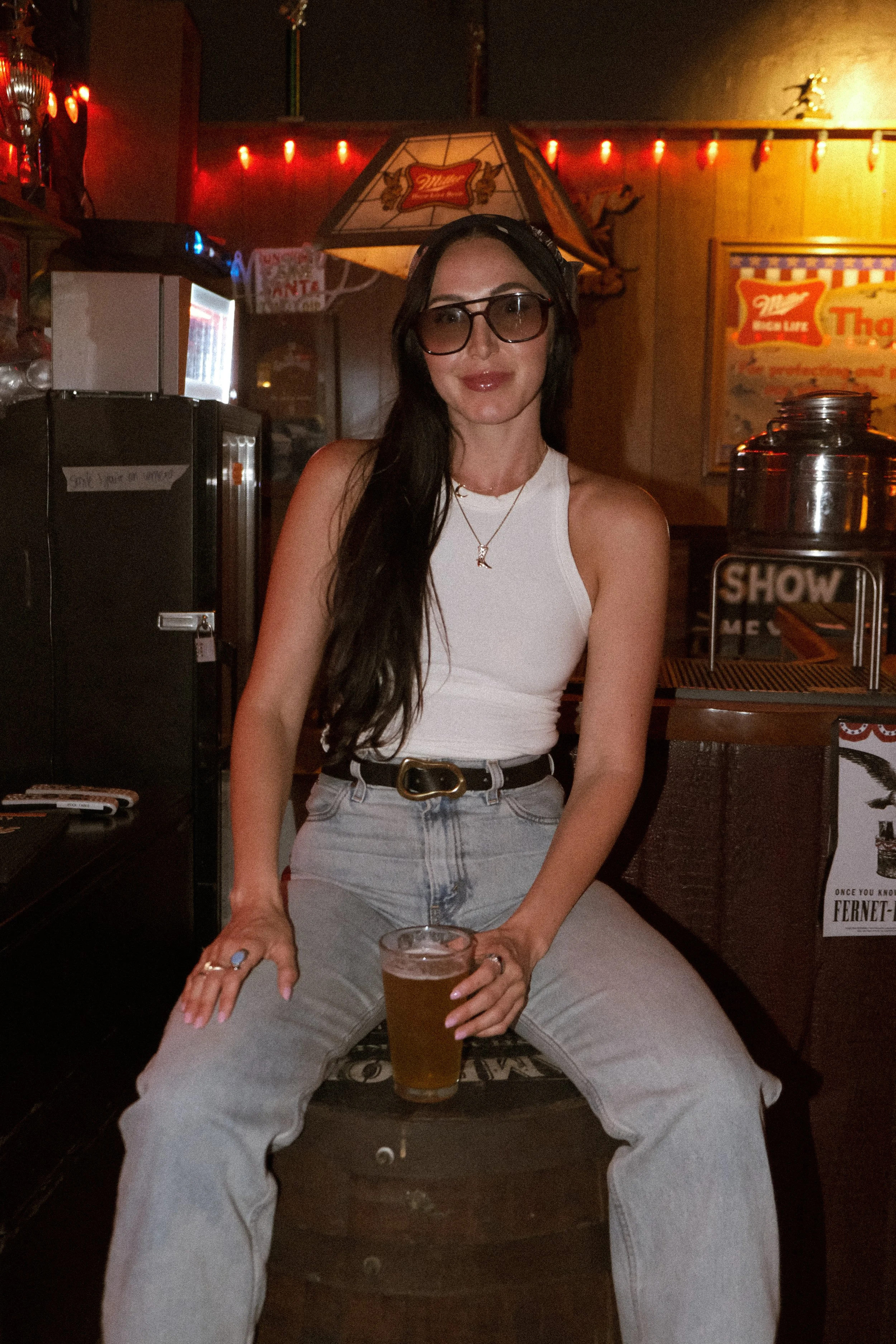 A woman with long dark hair wearing glasses, a white sleeveless top, and high-waisted light gray jeans, sitting on a wooden barrel in a bar or pub, holding a glass of beer, with dim lighting and neon signs in the background.