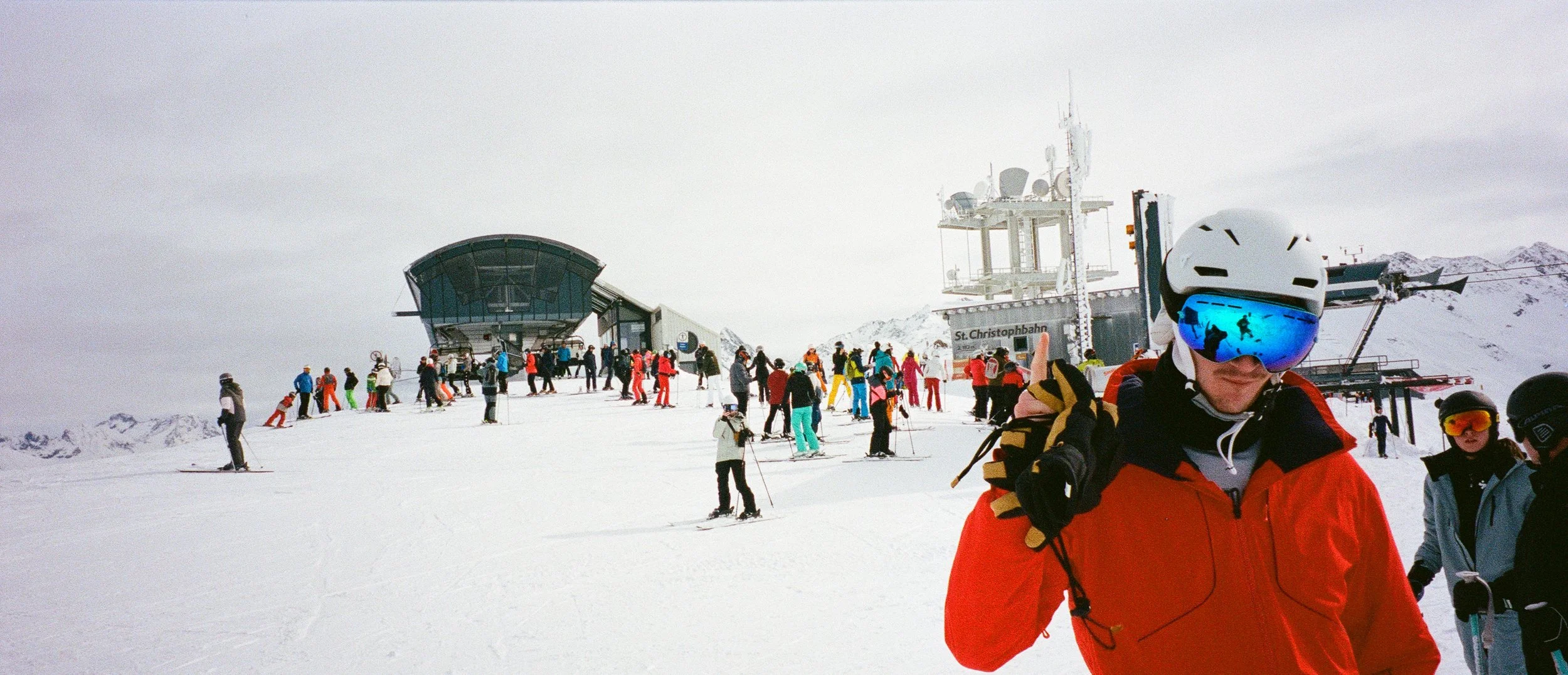 A busy ski resort with people skiing on snow and a person in the foreground wearing ski goggles, a white helmet, and an orange jacket.