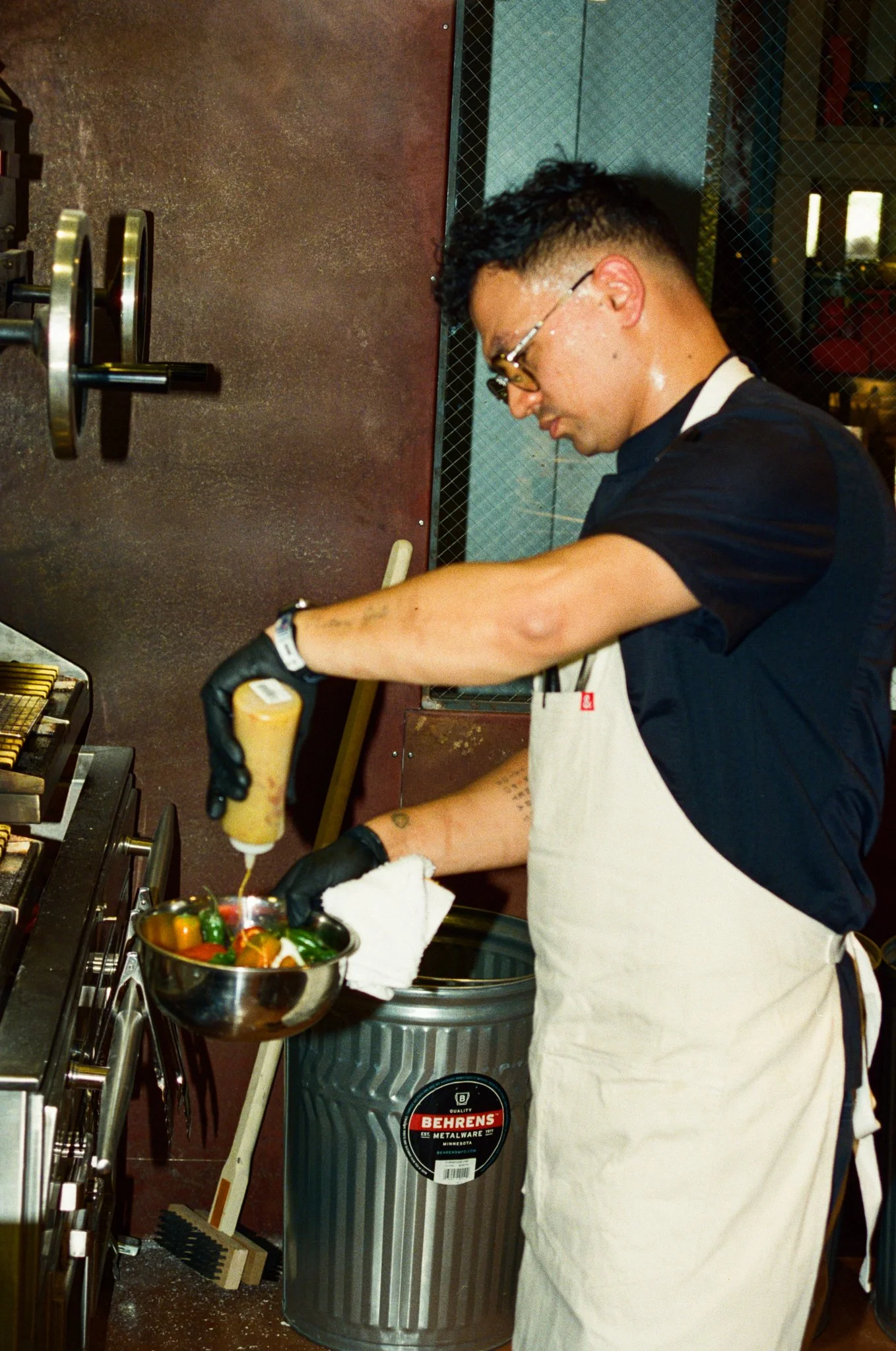 A man wearing glasses, a black shirt, and a beige apron is cooking with a squeeze bottle of sauce, pouring it into a bowl of vegetables in a kitchen.