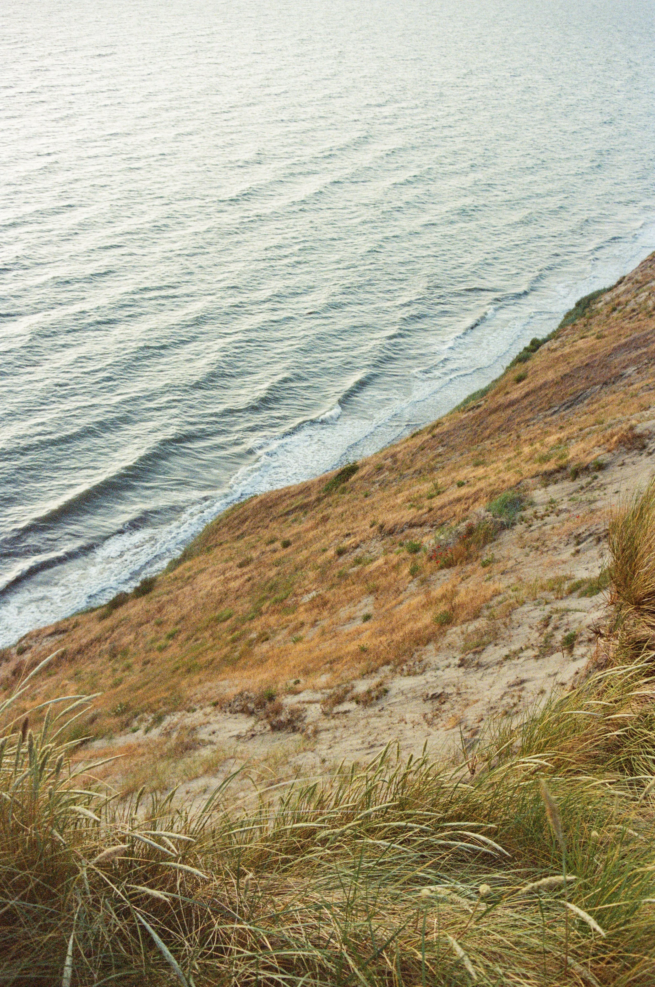 View of a sandy coastal hillside with tall grass, overlooking the ocean with small waves and a partially cloudy sky at sunset.