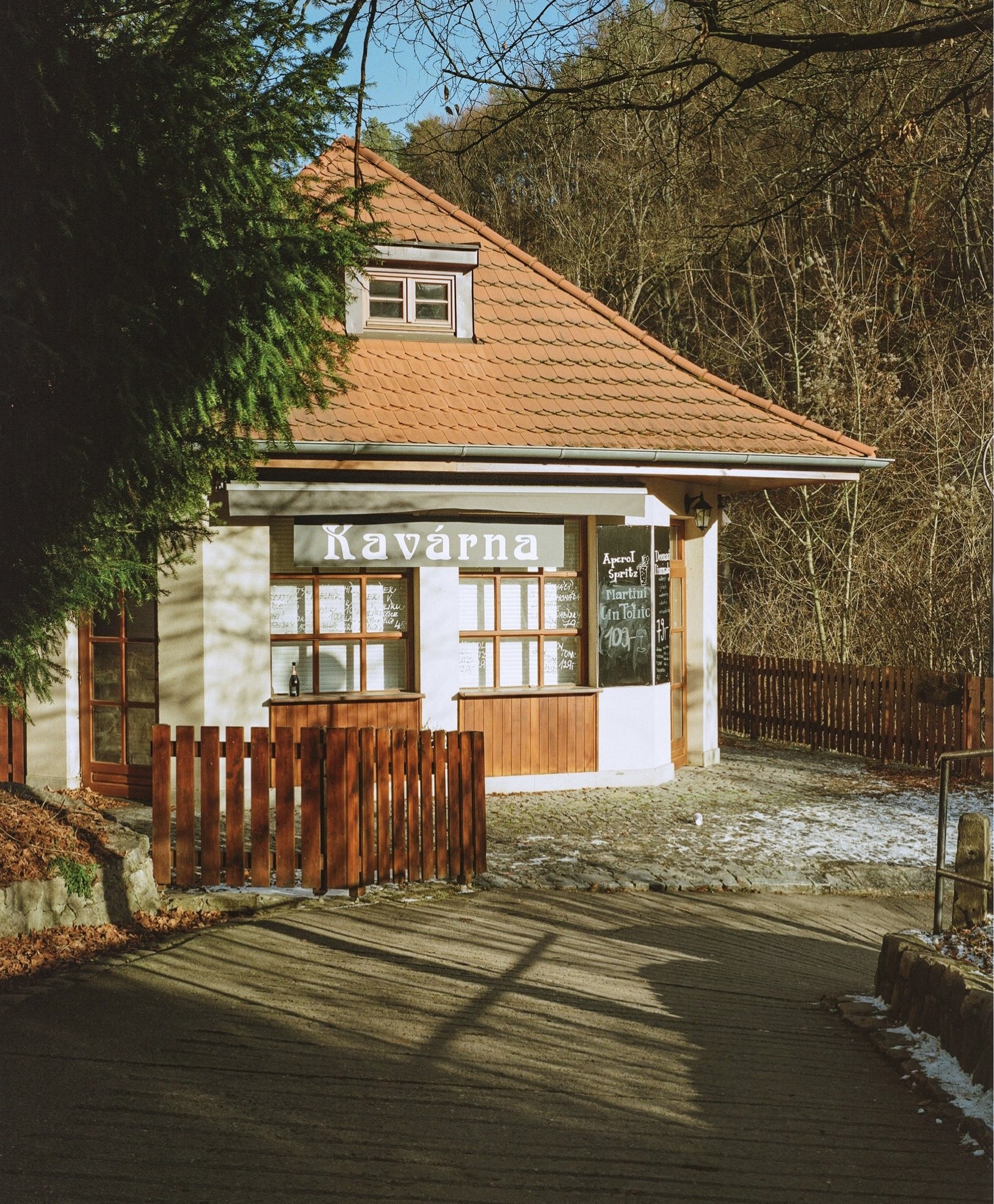 A small building with a sign that says "Kavárna" (Café) in Czech, featuring a red-tiled roof, wooden fences, large windows, and outdoor menu signs, surrounded by trees and a cobblestone path.