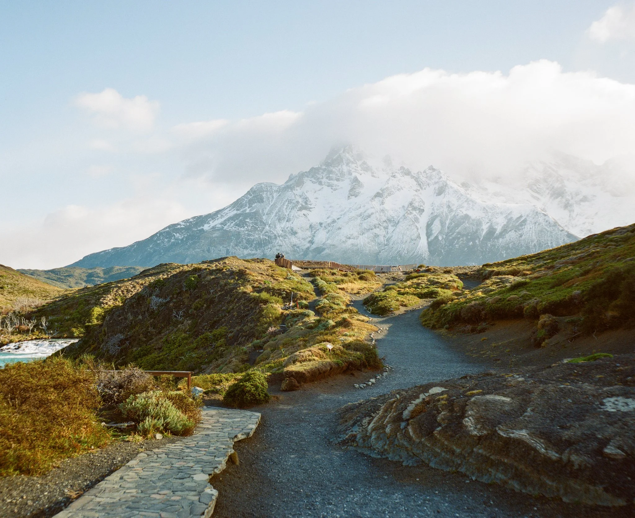 A scenic mountain landscape with a gravel and stone pathway winding through green hills towards snow-capped peaks in the distance.