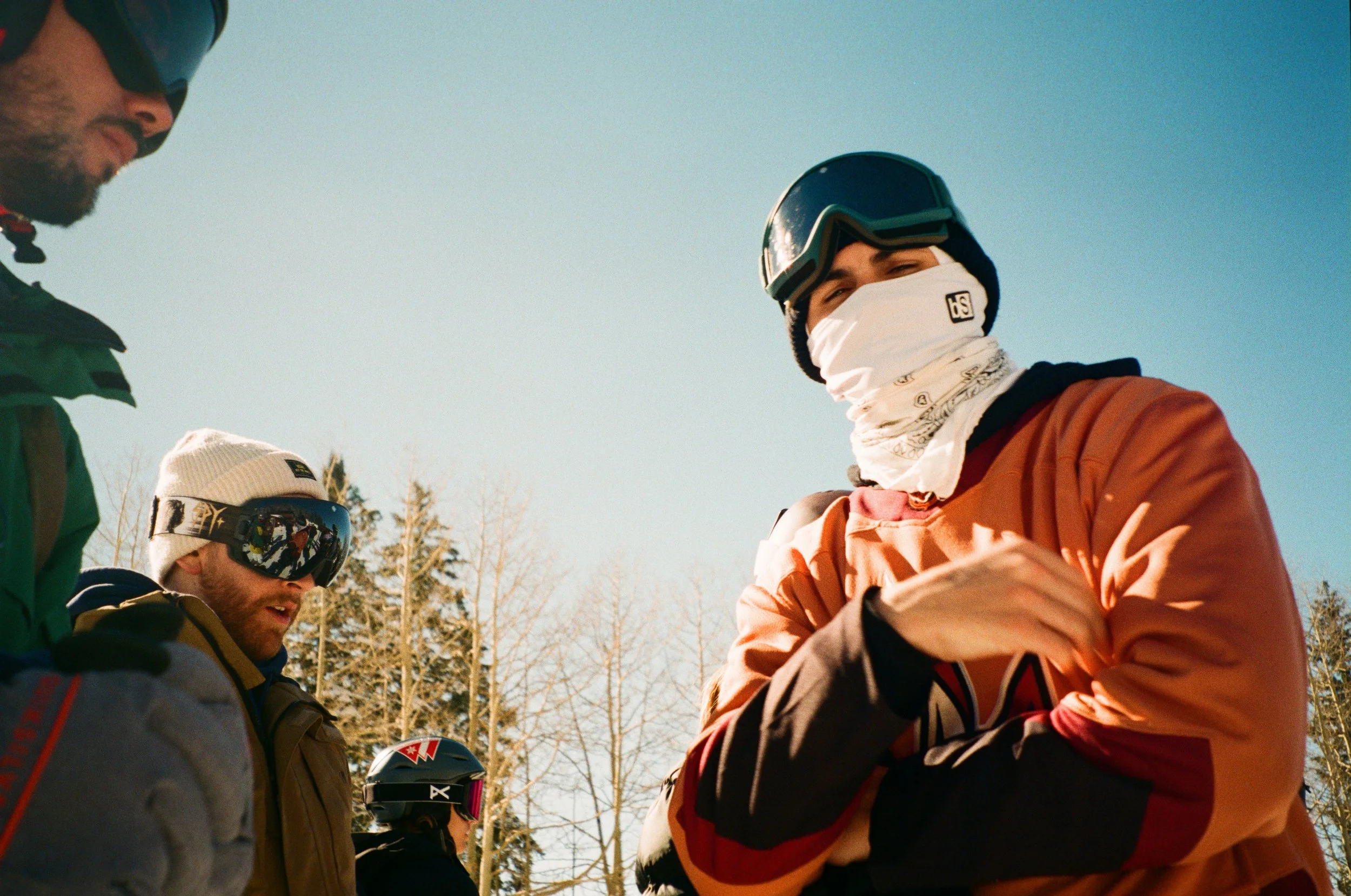 Group of snowboarders on a snowy mountain outdoors, wearing winter gear including helmets, goggles, and face masks, with a background of bare trees and a clear blue sky.