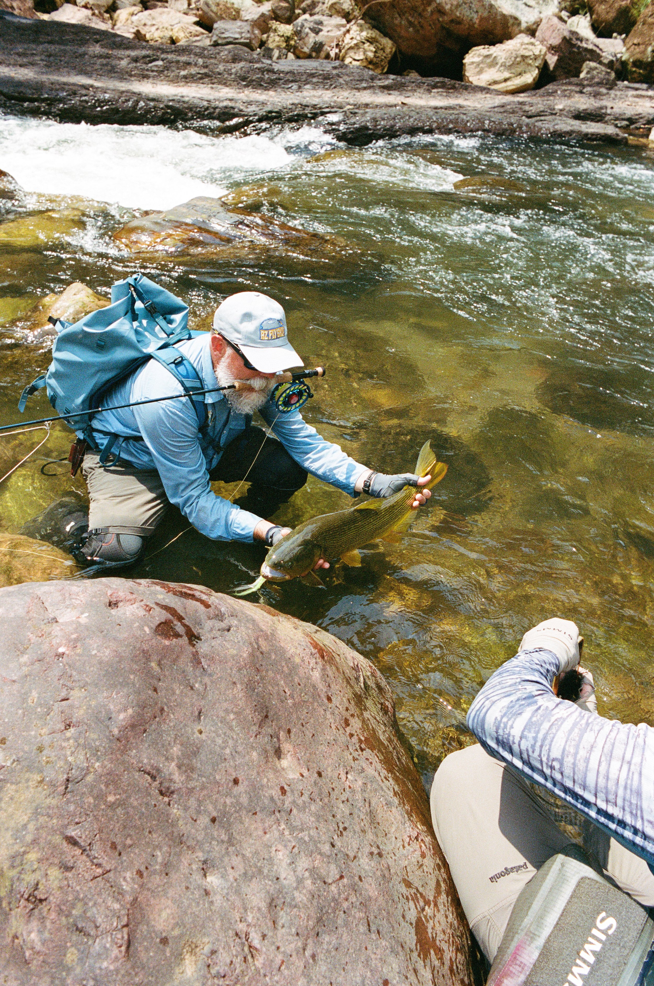 A man with a beard, wearing outdoor gear, kneeling on rocks in a river, holding a fish he has caught.
