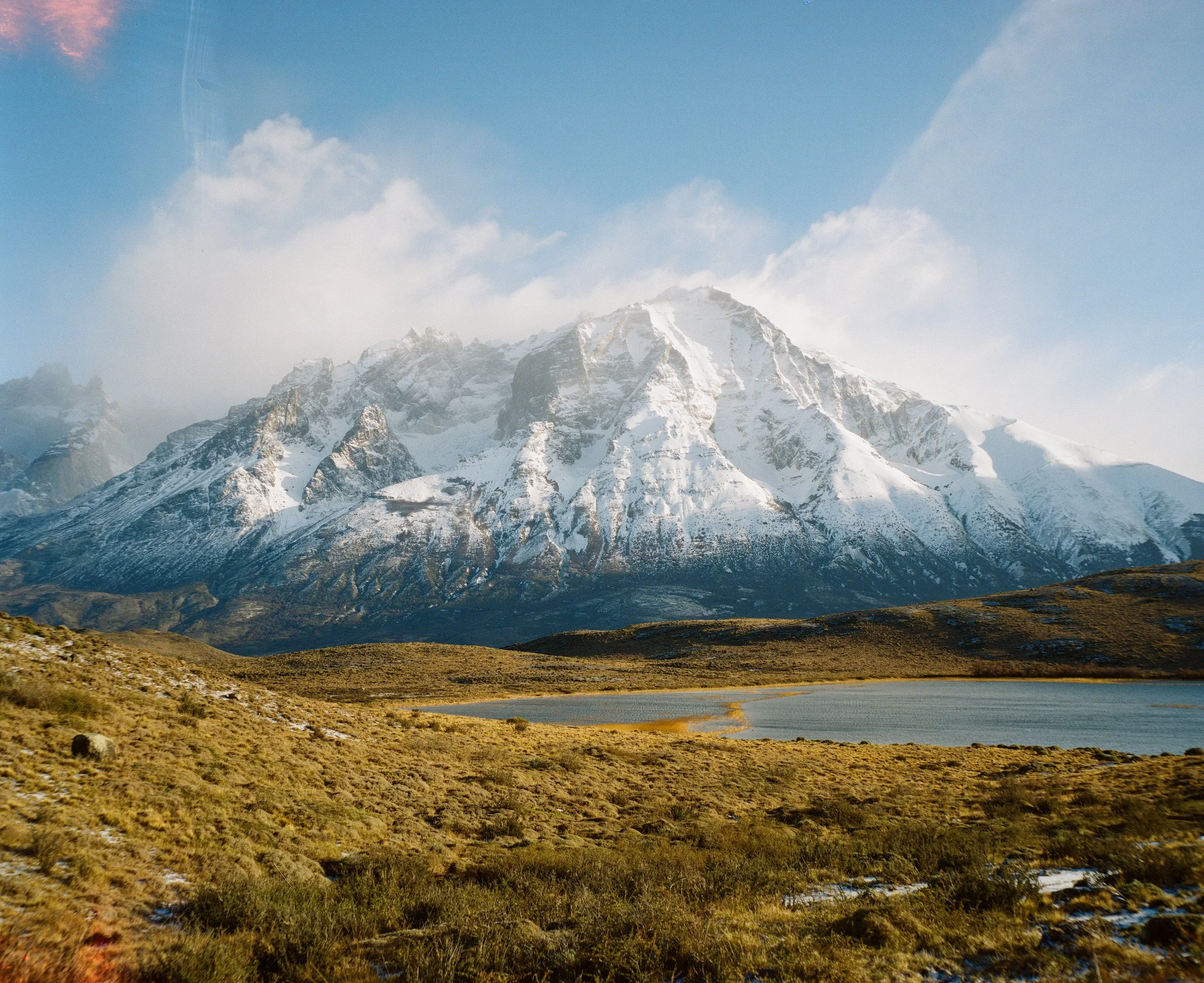 Snow-capped mountain range reflecting in a lake surrounded by grassy plains under a cloudy sky.