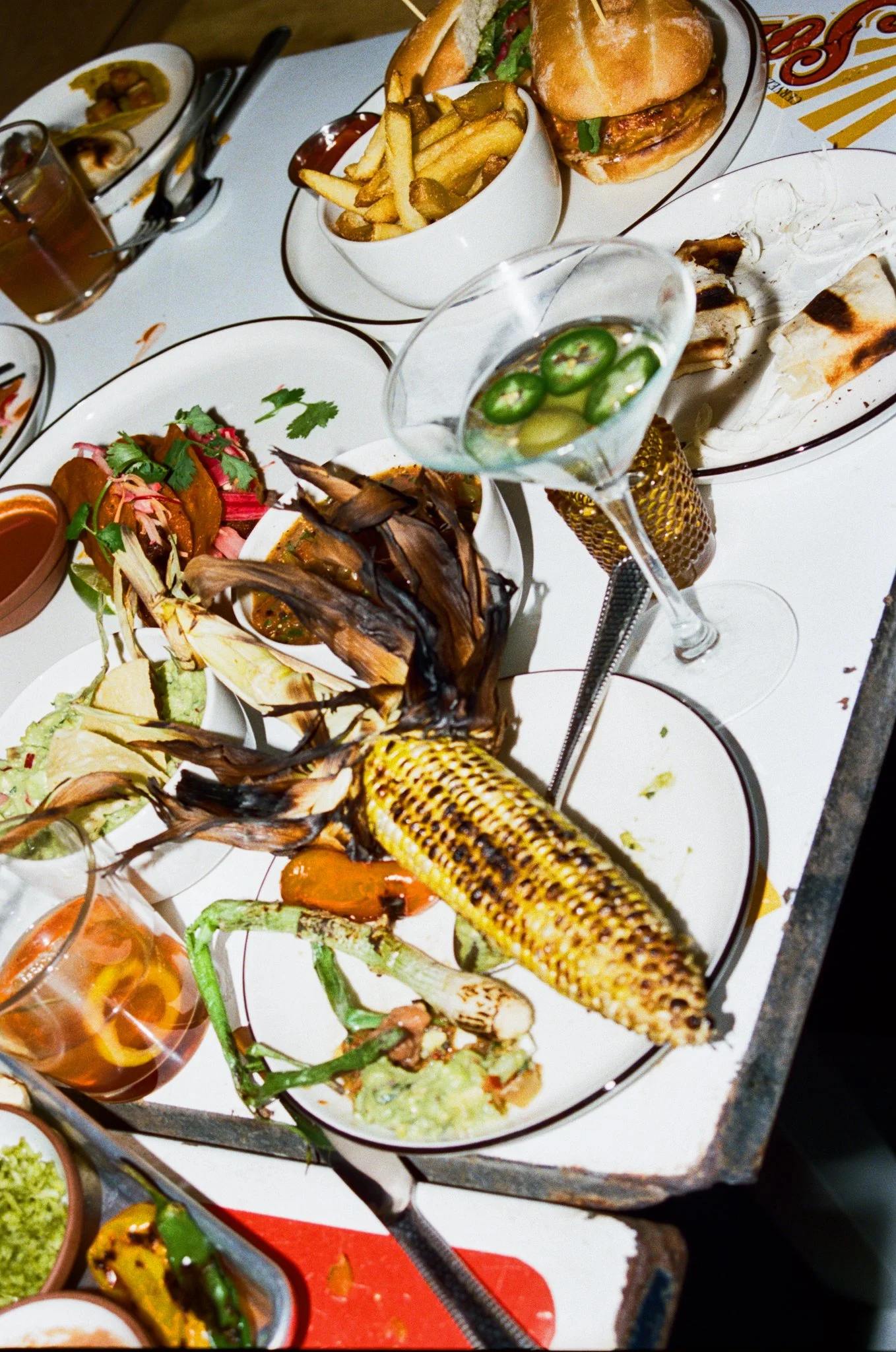 A table filled with various dishes including grilled corn on the cob, grilled vegetables, a burger with lettuce and tomato, a bowl of French fries, and several glasses of beverages, one with jalapeño slices.