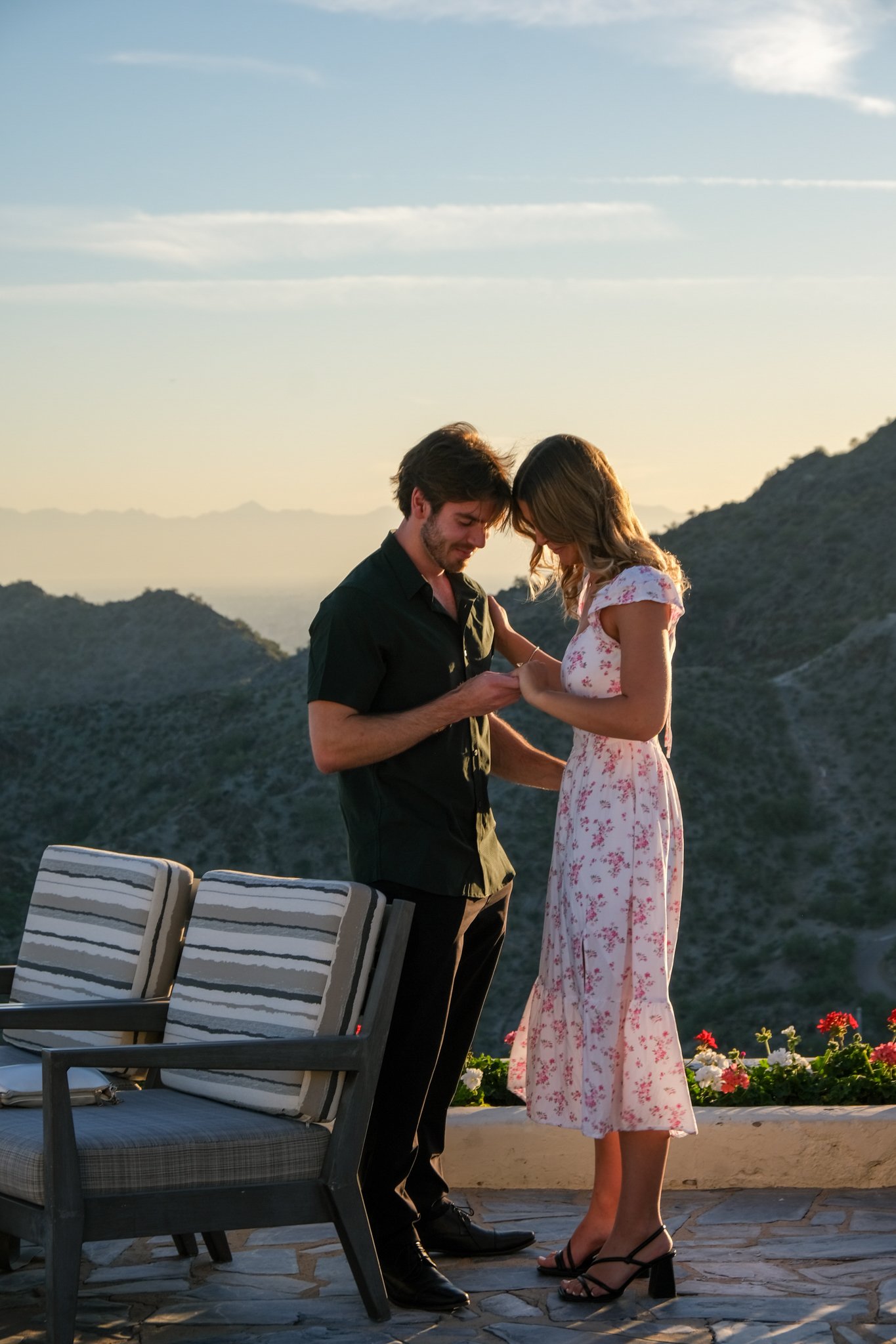A couple standing on a patio during sunset, with mountains in the background. The man is wearing a black shirt, and the woman is wearing a light pink floral dress and heels. They appear to be sharing a romantic moment.