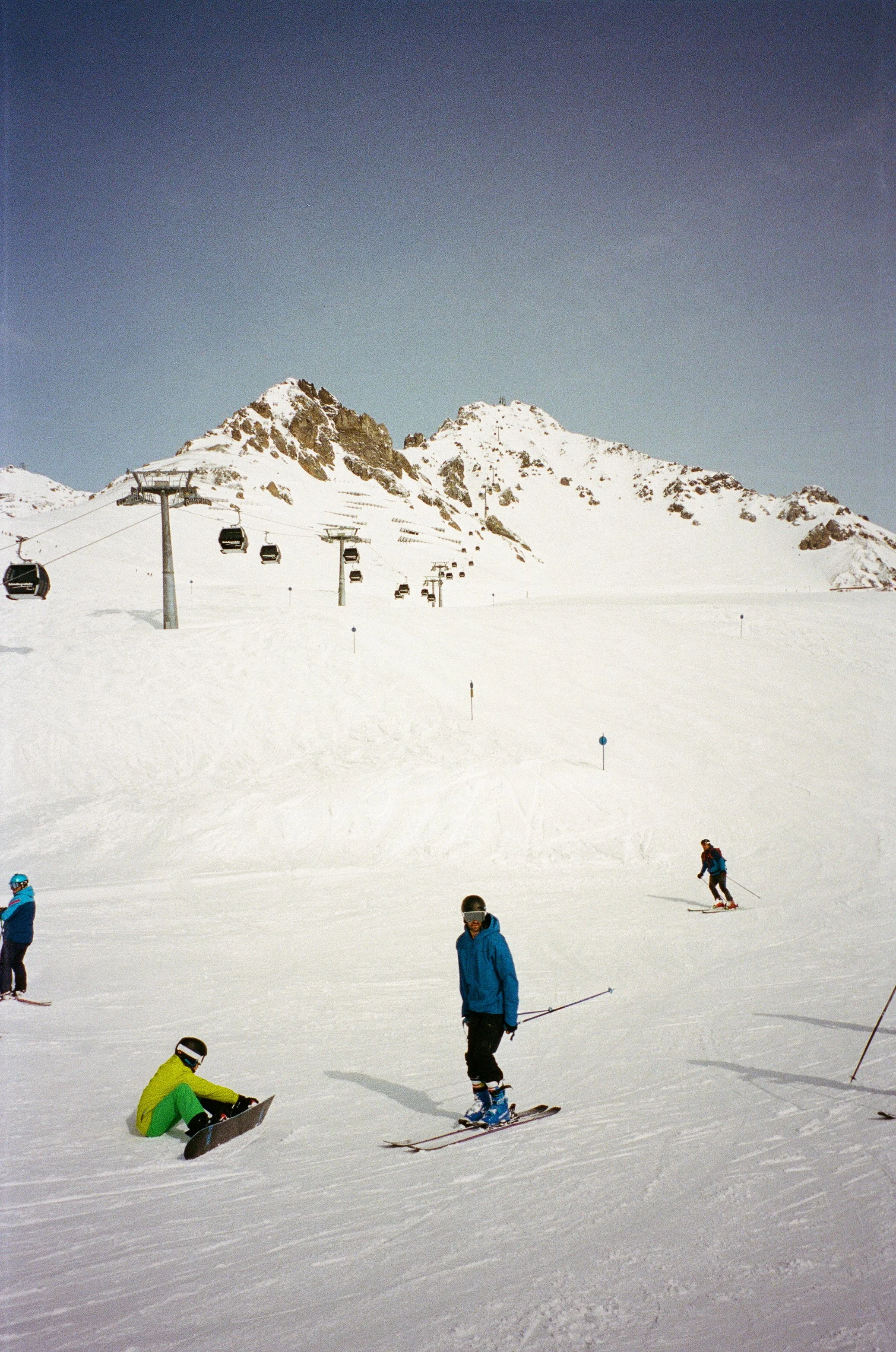 Skiers and snowboarders on a snowy mountain slope near ski lifts with mountain peaks in the background under a clear sky.