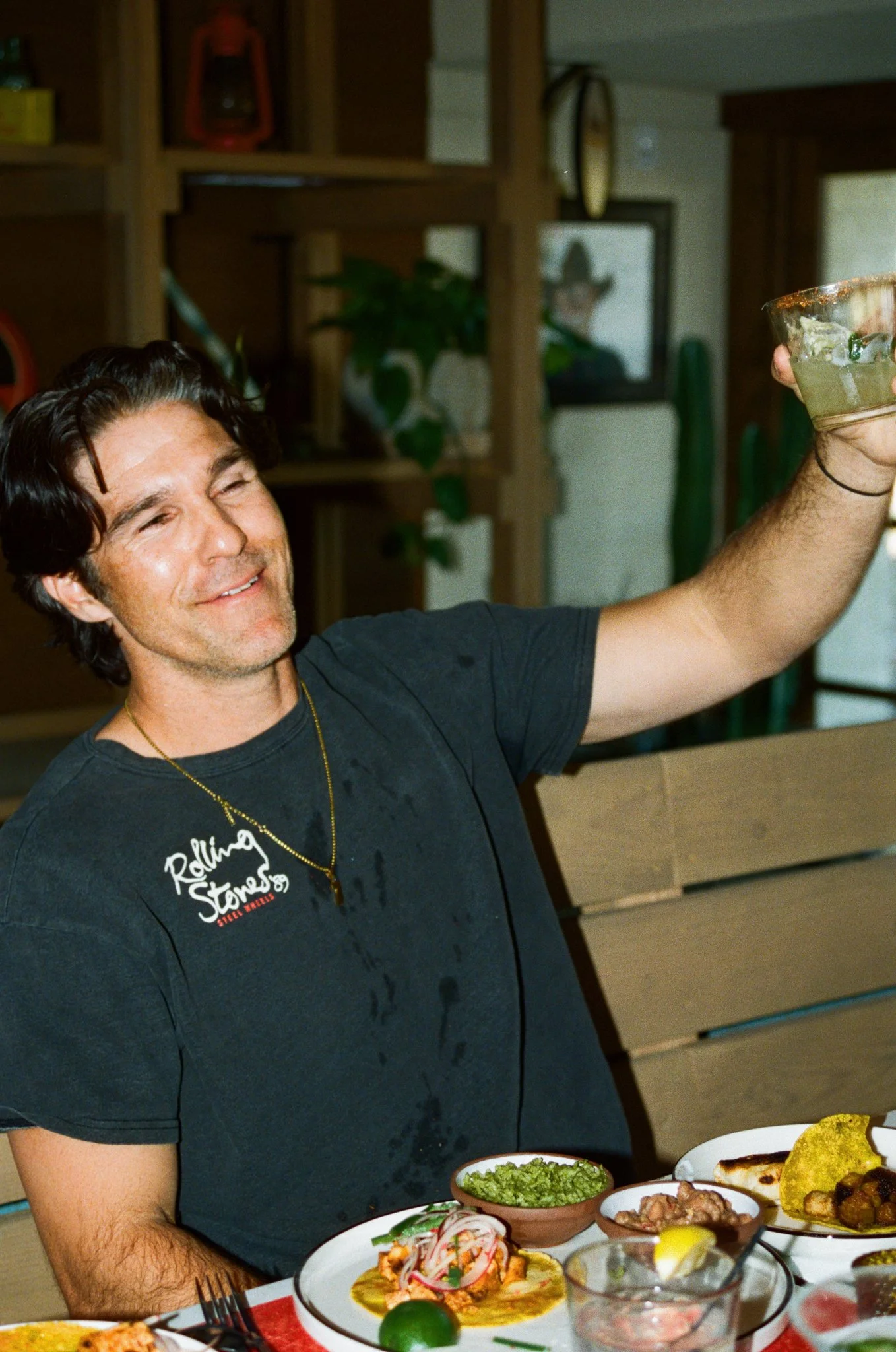 A man with dark hair, smiling, raising a glass of margarita. He's wearing a black T-shirt with 'Rolling Stones' written on it, sitting at a table with Mexican food.