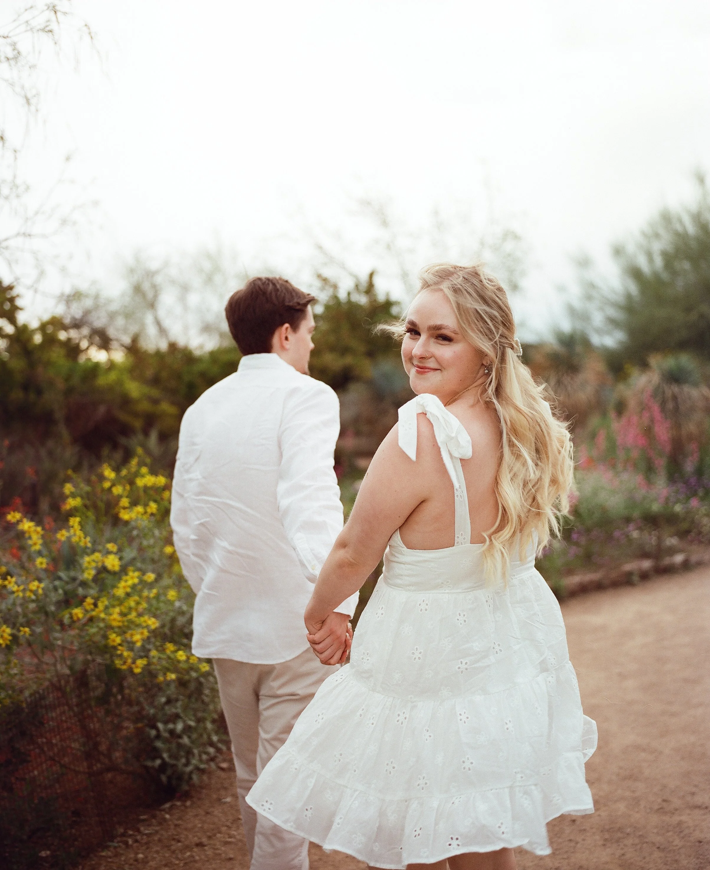 A young woman in a white dress holding hands with a young man in white shirt and light pants, standing outdoors in a garden with yellow and pink flowers, the woman standing and smiling at the camera.