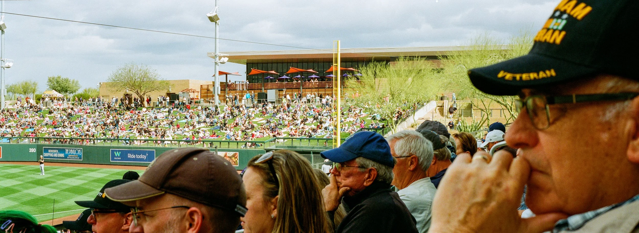 Crowd of spectators at a baseball game with a view of the field and a large seating area in the background, some wearing hats and glasses, with a cloudy sky above.