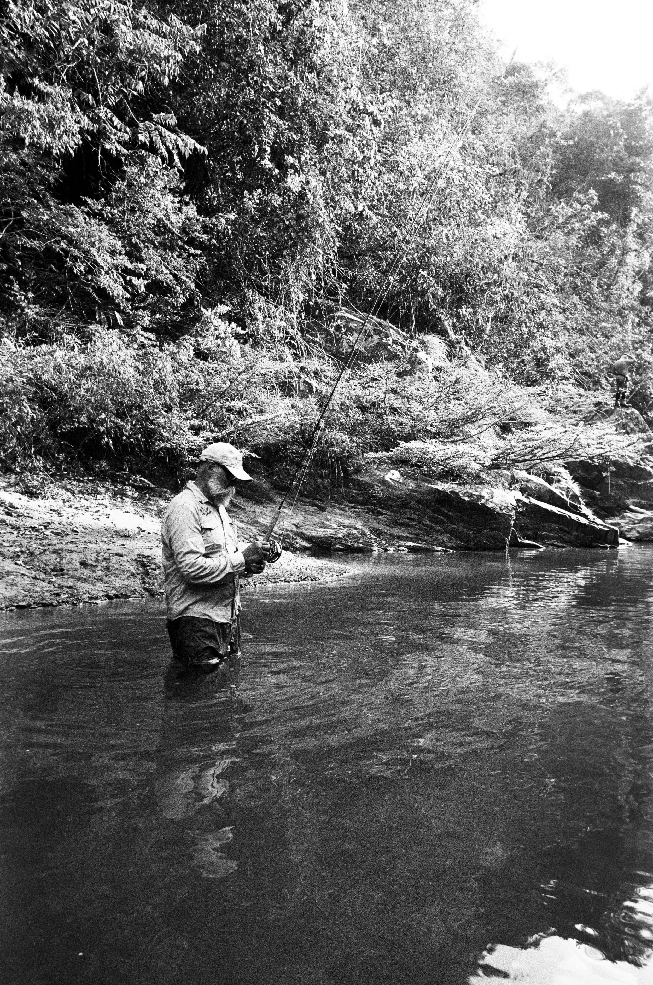 A person fishing in a river with dense trees and rocks in the background.