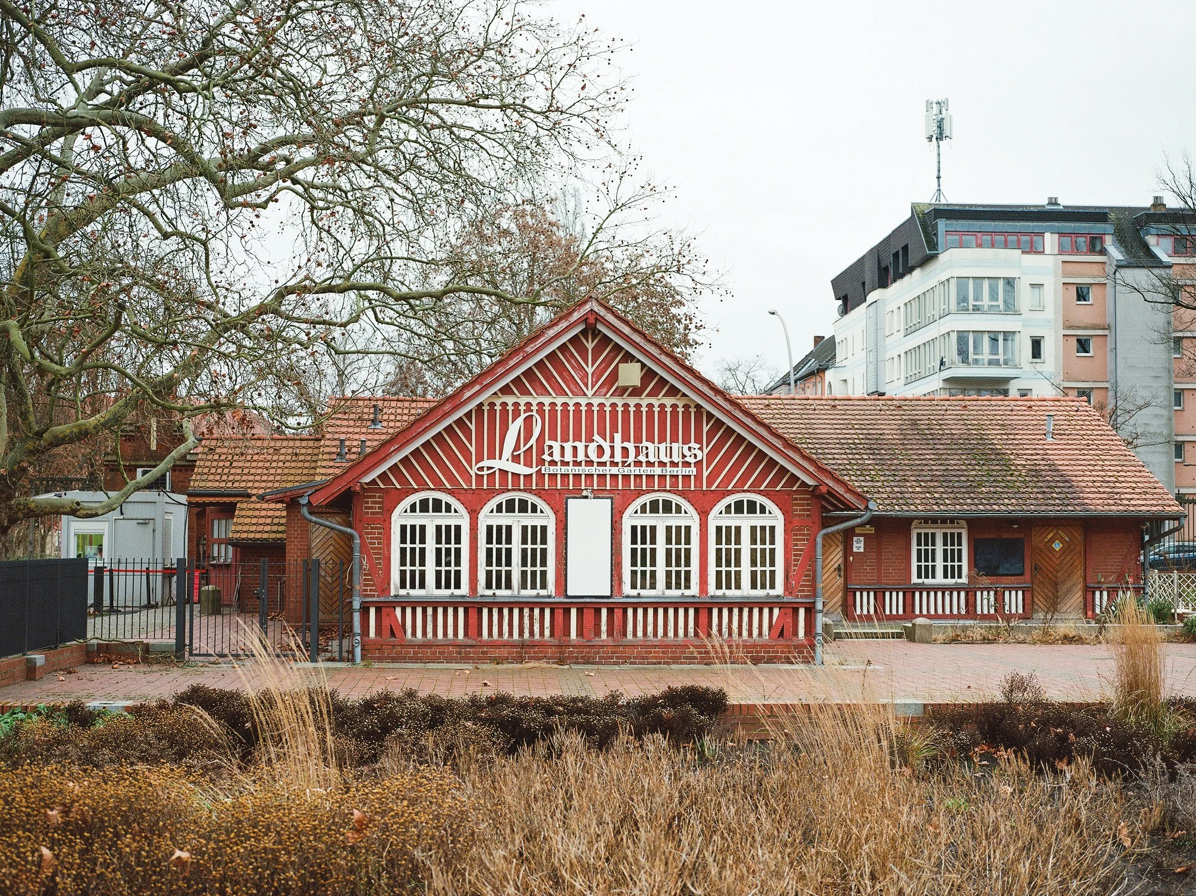 Red brick building with white-framed arched windows and a sign that reads "Landsäng" in Berlin, Germany, with leafless trees and modern apartment buildings in the background.