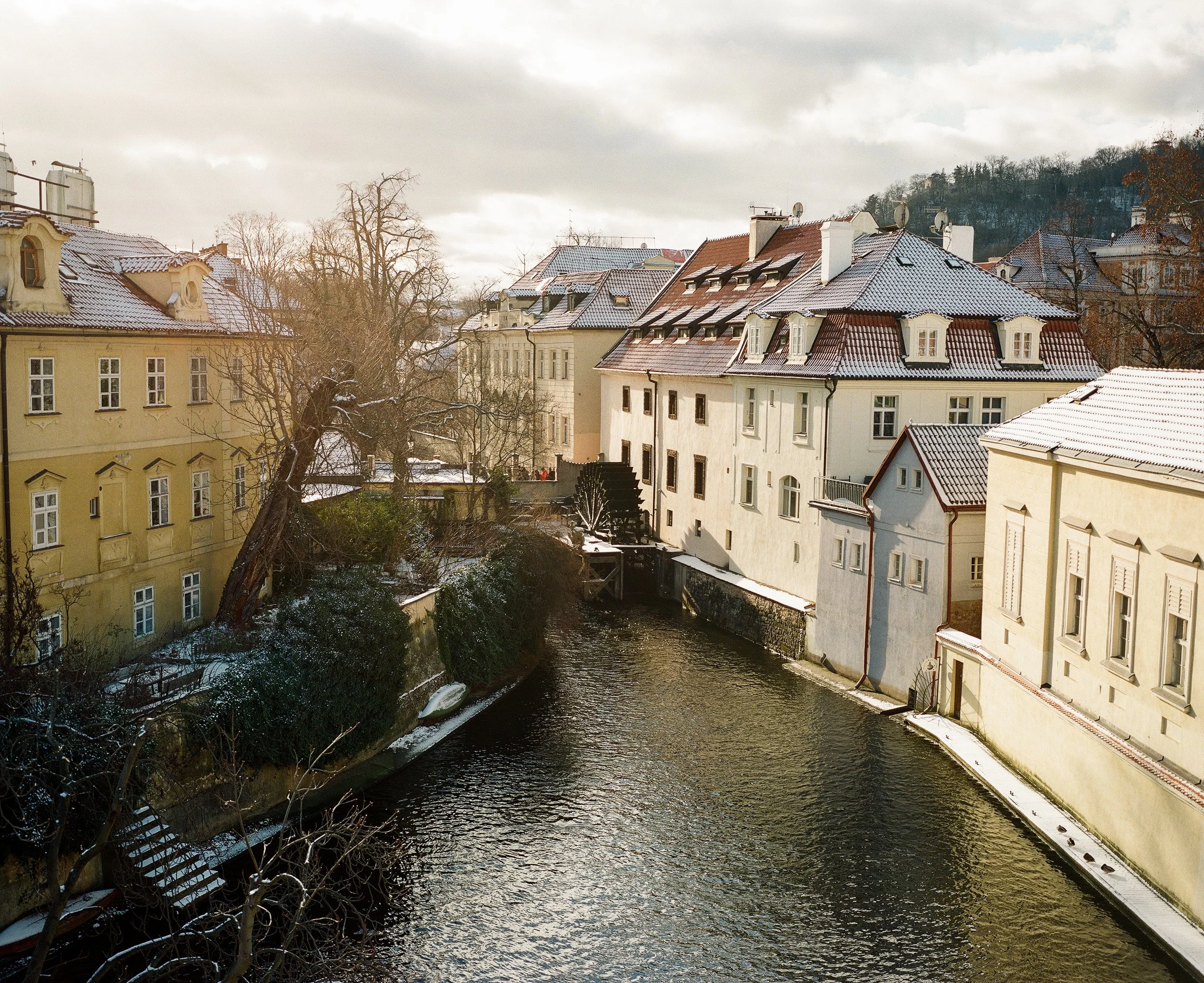 A canal lined with historic buildings with snow-covered rooftops on a winter day, with clouds in the sky and a distant hill in the background.