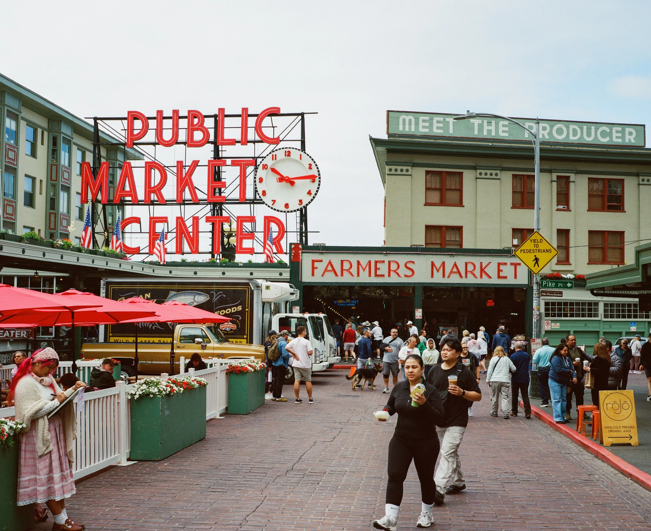 People walking and shopping at Pike Place Market in Seattle, Washington, with signs reading "Public Market Center," "Farmers Market," and "Meet the Producer."