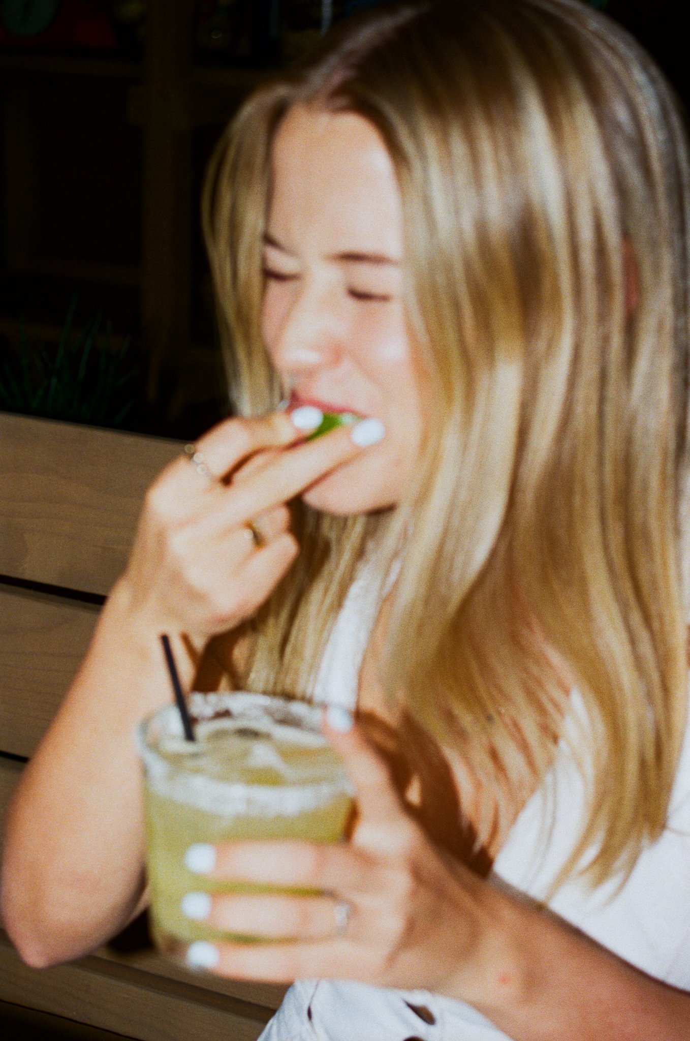 A woman with long blonde hair is smiling while eating a slice of lime from her hand, holding a cold drink with a straw in her other hand.