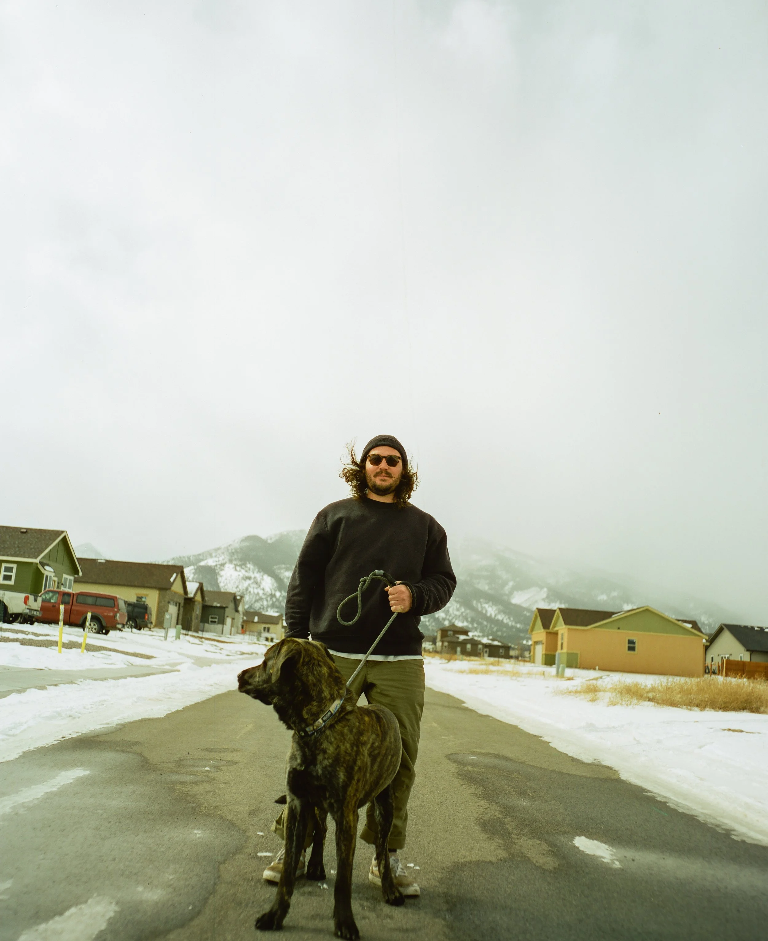 A man with long hair, sunglasses, a gray beanie, and a black sweatshirt standing on a snowy street holding a large brindle dog on a leash, with snow-covered mountains and houses in the background.
