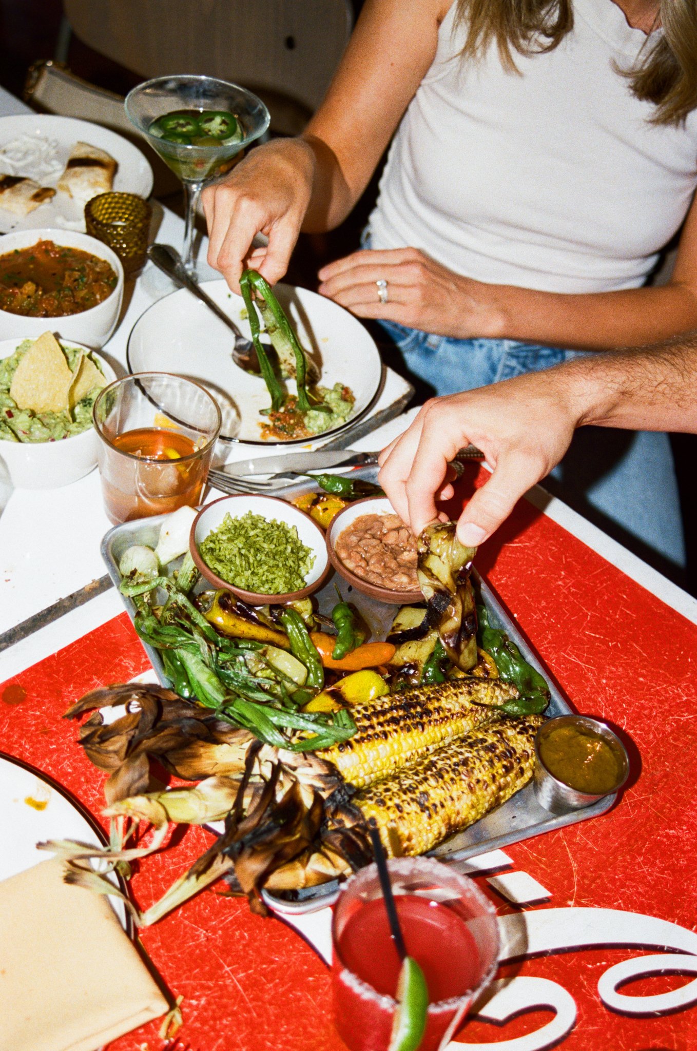 People sharing a Mexican meal with grilled corn, salsa, and guacamole, with drinks on a red table.