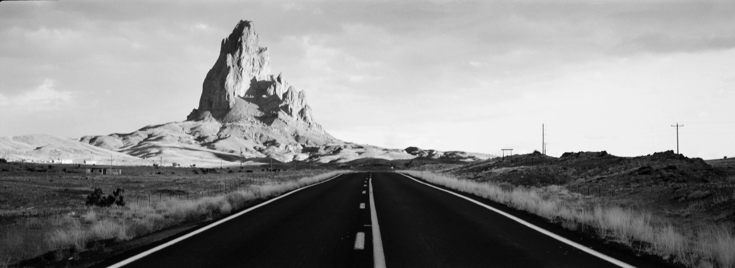 Black and white photo of a straight road leading towards a rocky mountain in a barren landscape with scattered small buildings and power lines.