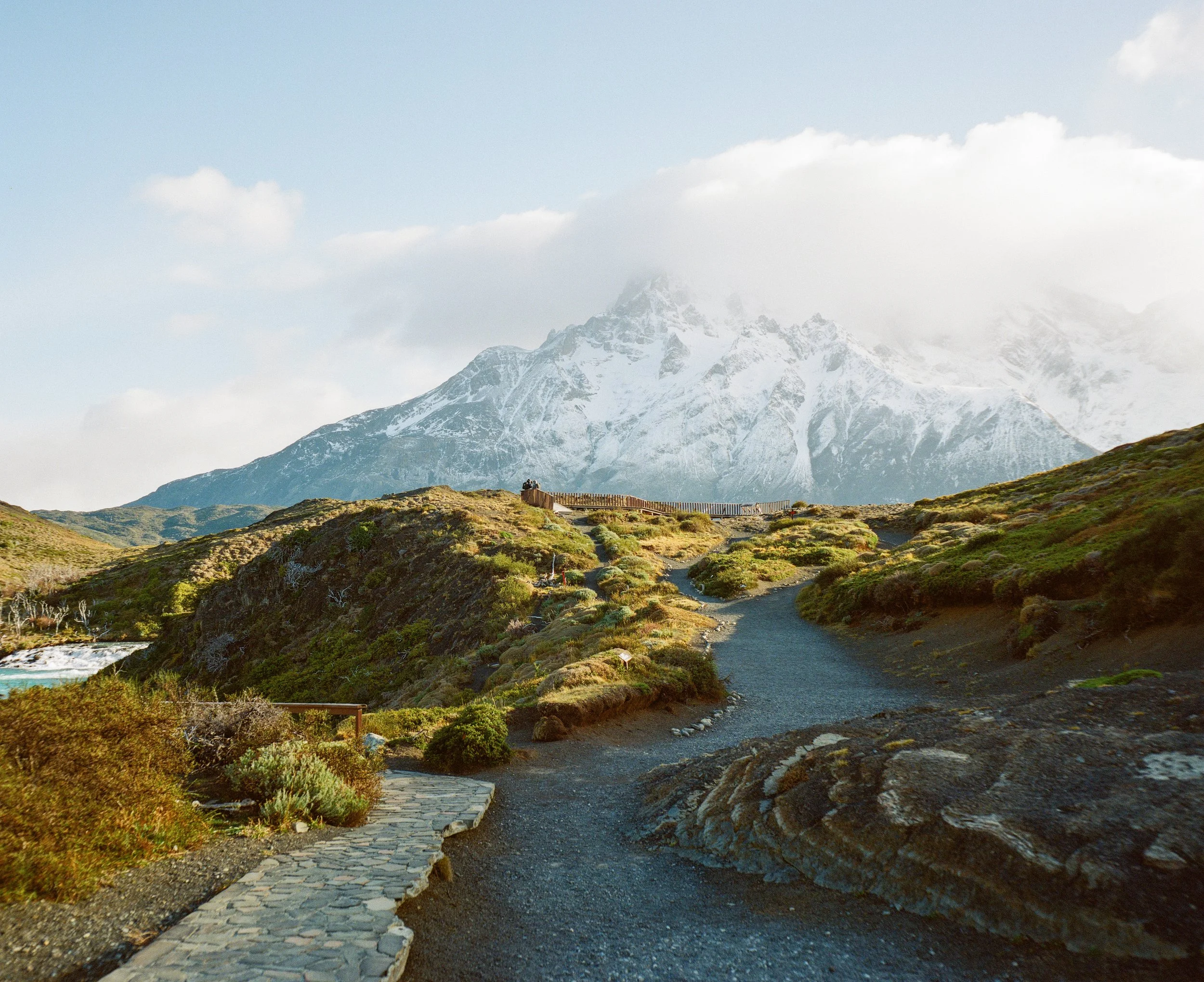 A rugged trail in a mountainous landscape with snow-capped peaks in the background. A stone-lined path leads upward, surrounded by patches of green and brown vegetation. A viewing platform with a few people is visible near the top of the trail.
